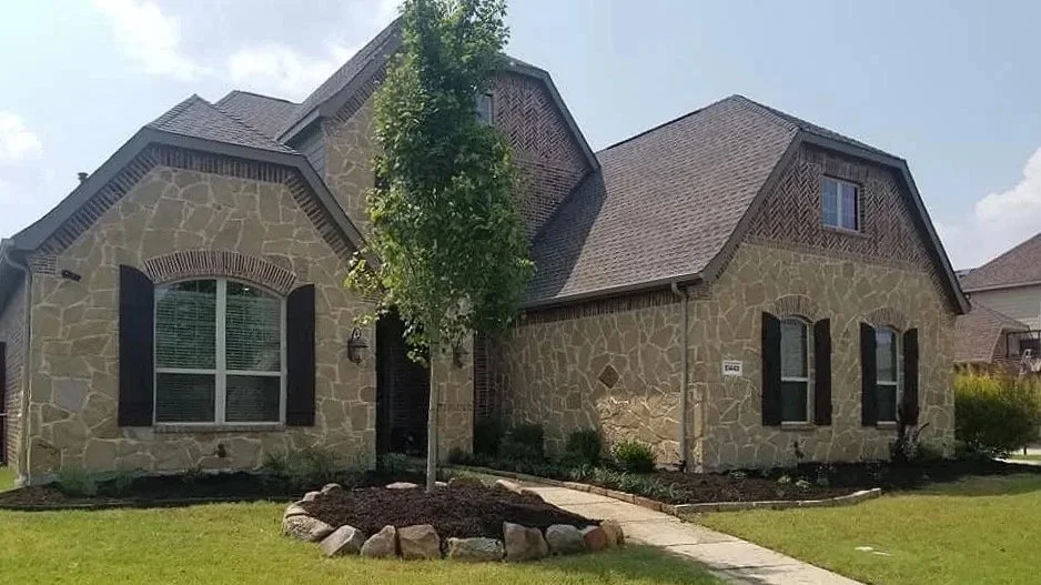 Front view of a house with stone walls, two front windows with black shutters, a small tree, and a lawn.