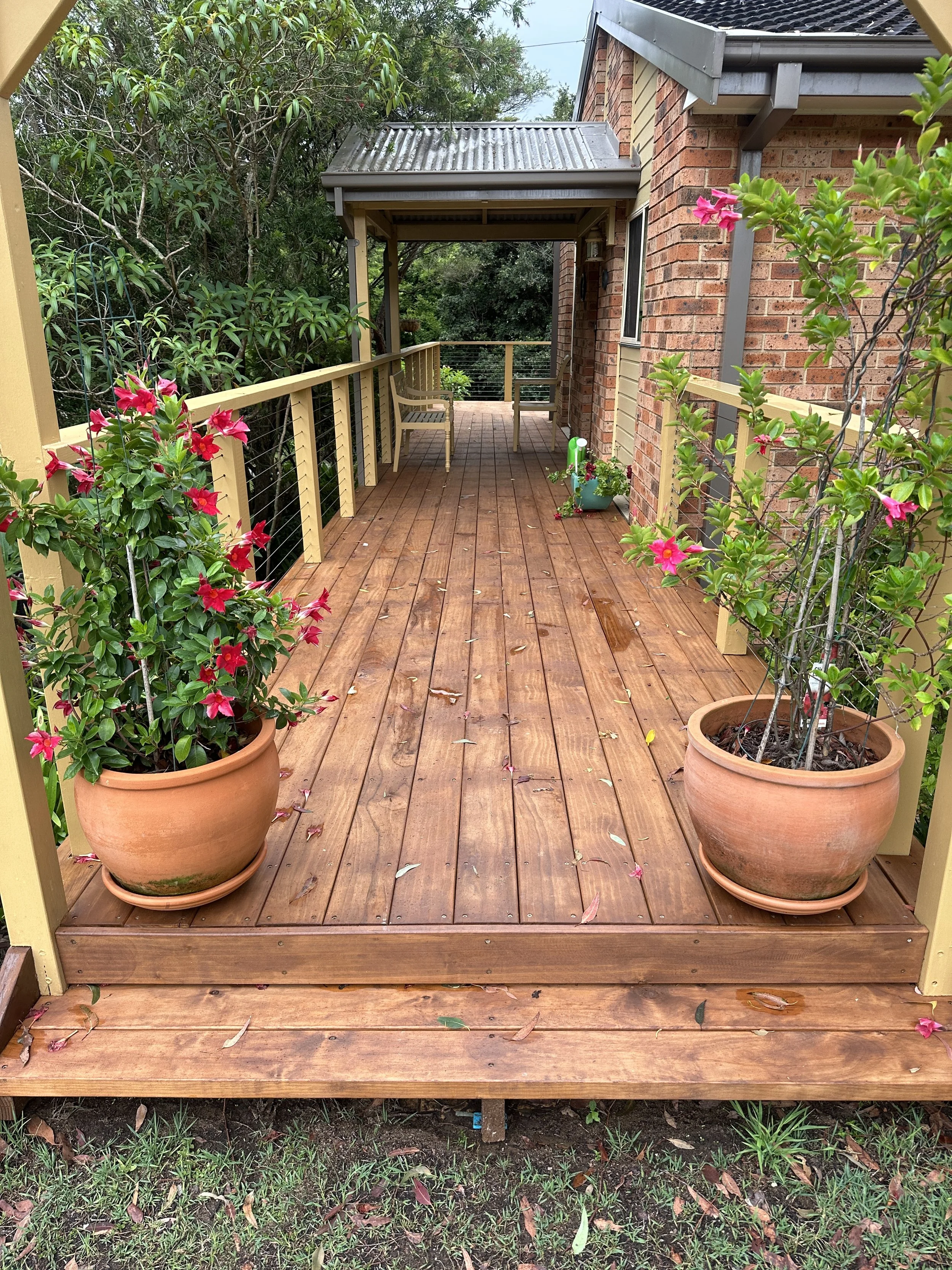A wooden porch attached to a brick house, decorated with potted plants and surrounded by greenery. The porch features a small roof, a bench, and a railing, with some fallen leaves on the wooden floor.