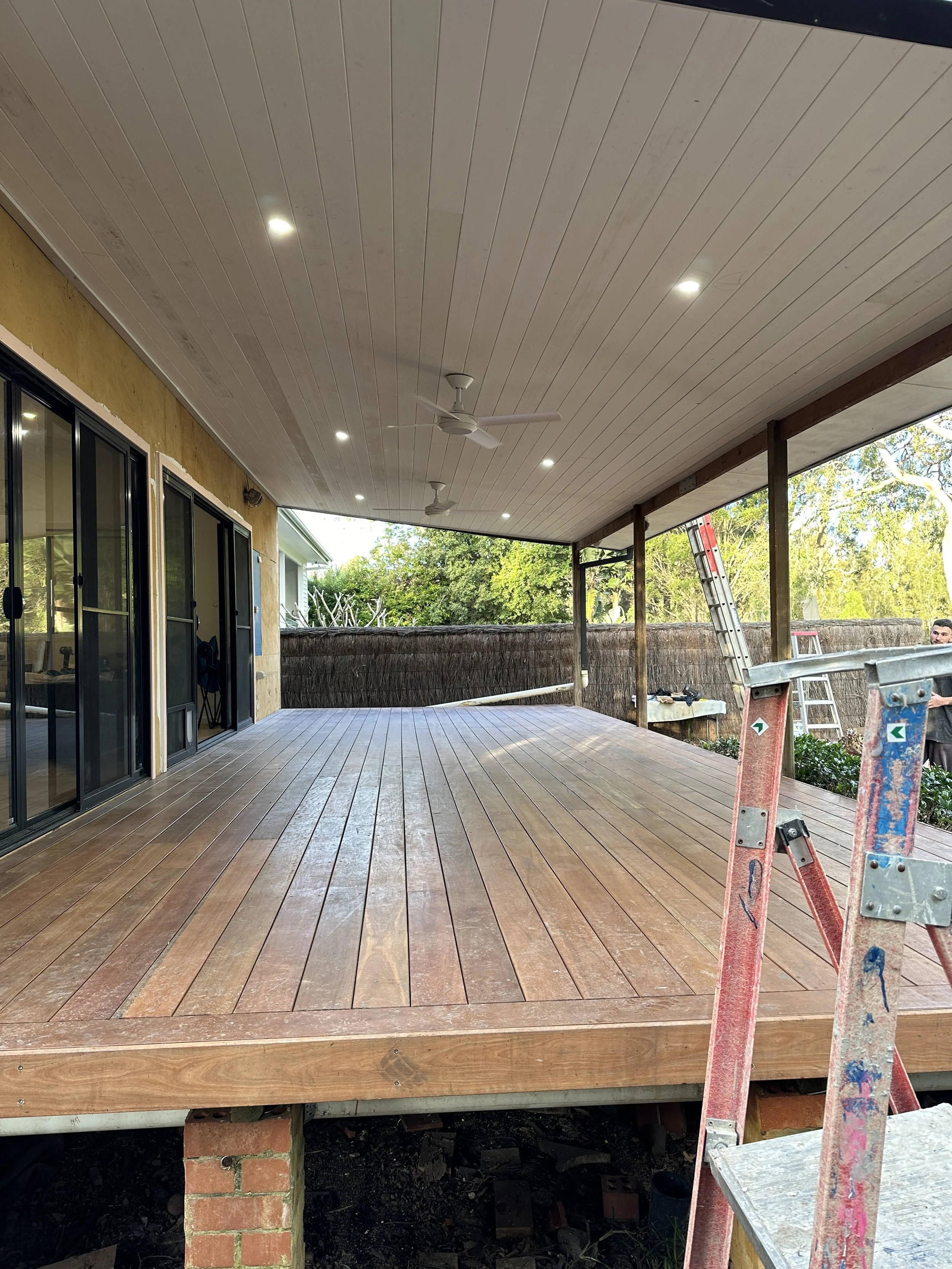 A covered wooden patio with ceiling fans and recessed lighting, adjacent to a house with sliding glass doors, overlooking a green yard with trees and a fence, in the process of being built or renovated.