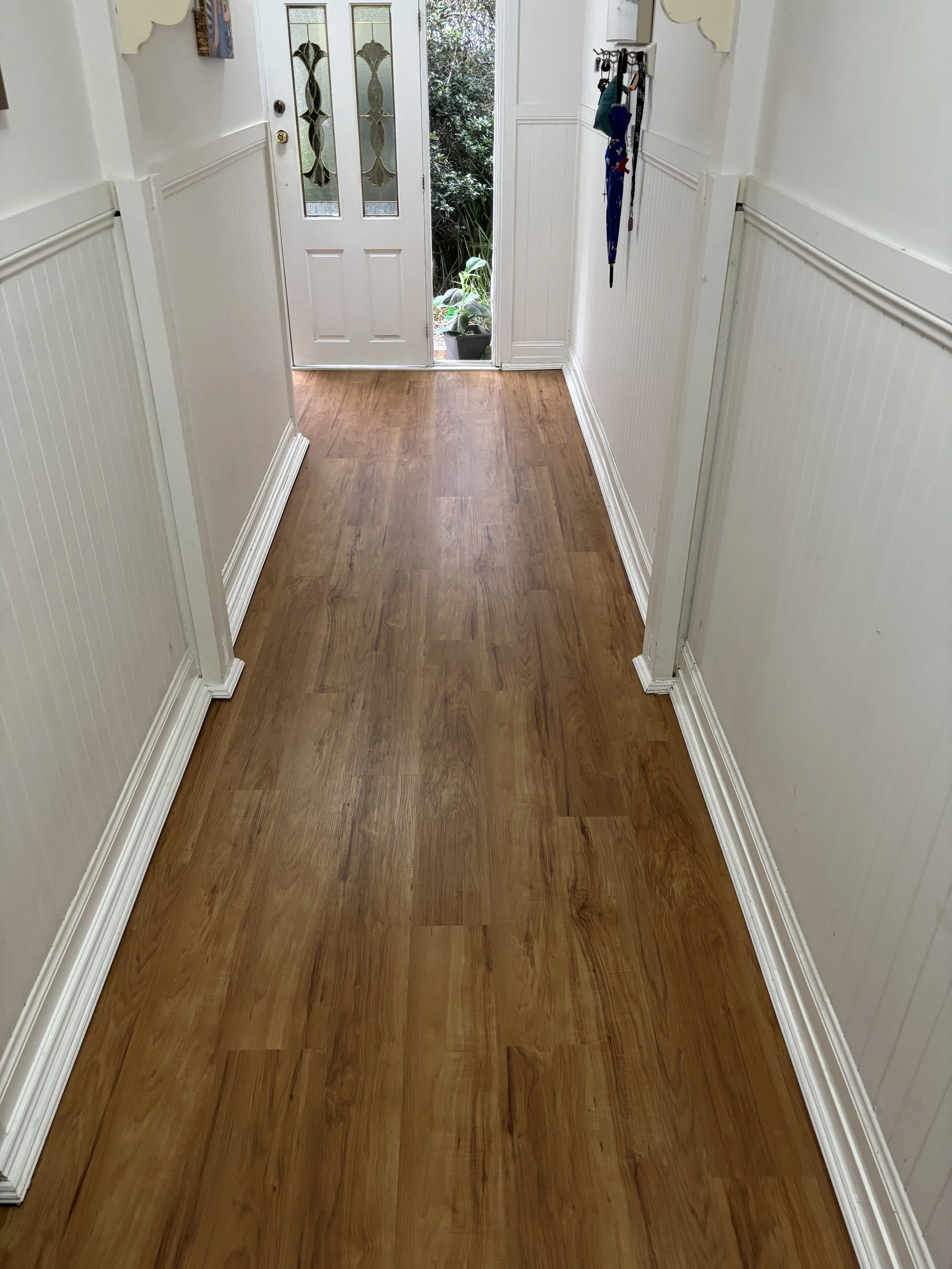 A hallway with hardwood floor and white walls. At the end, a white door with decorative glass panels and a screen door. Outside, plants and greenery are visible through the door.