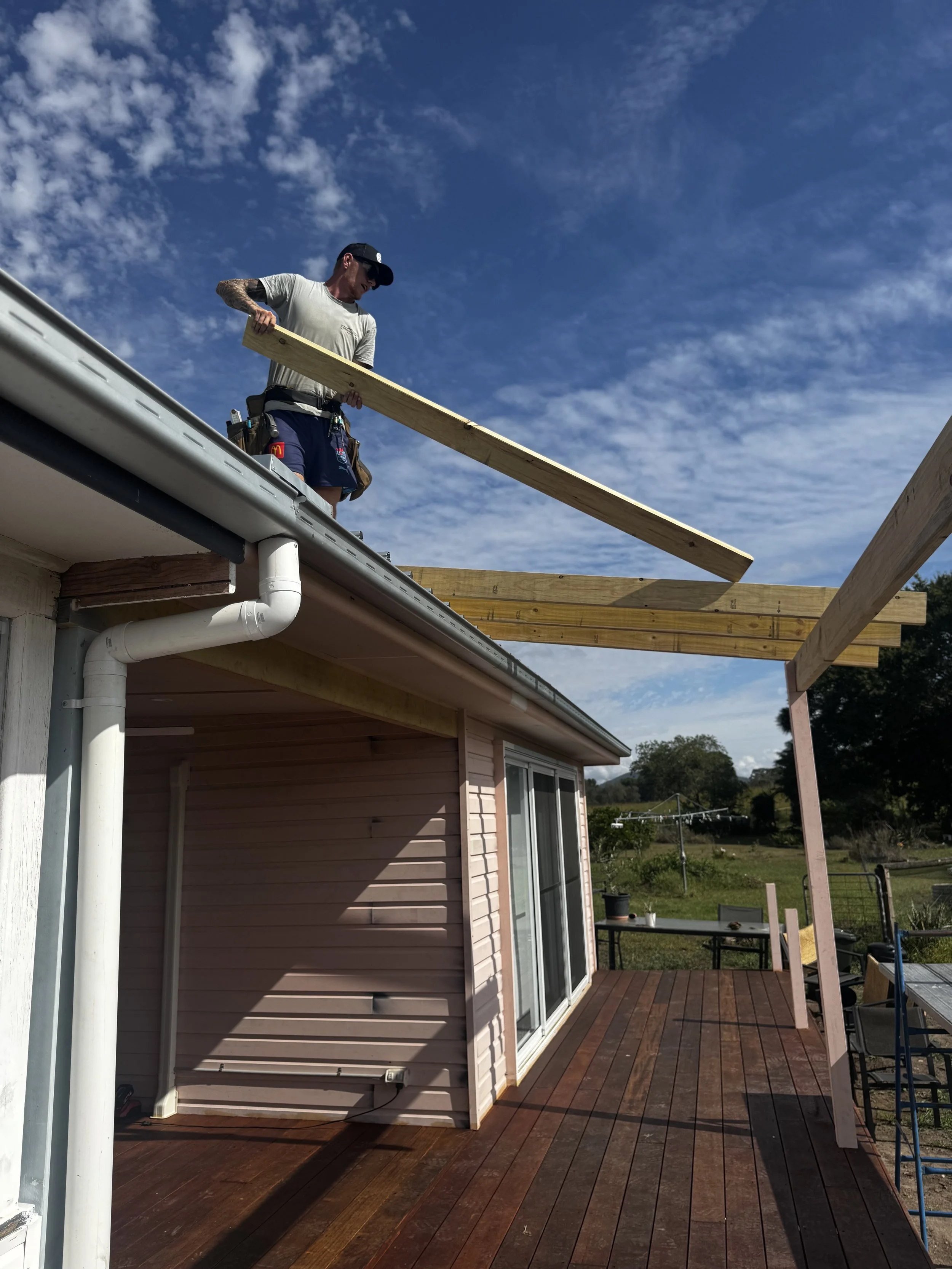 An individual in construction gear working on a wooden deck extension outside a house, under a bright blue sky with some clouds.