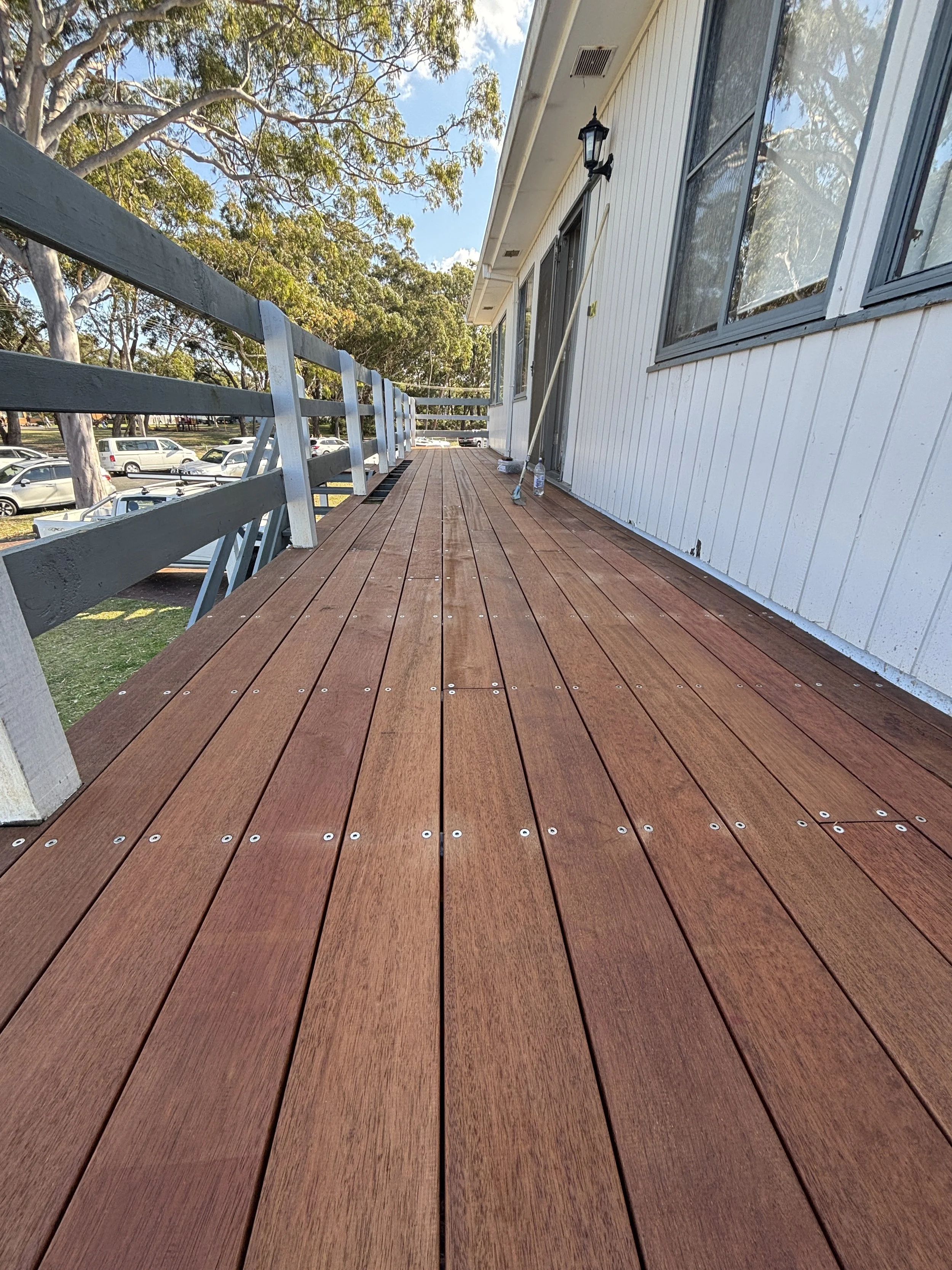 Wooden deck with metal railing attached to a white house, with trees and parked cars visible in the background on a clear sunny day.