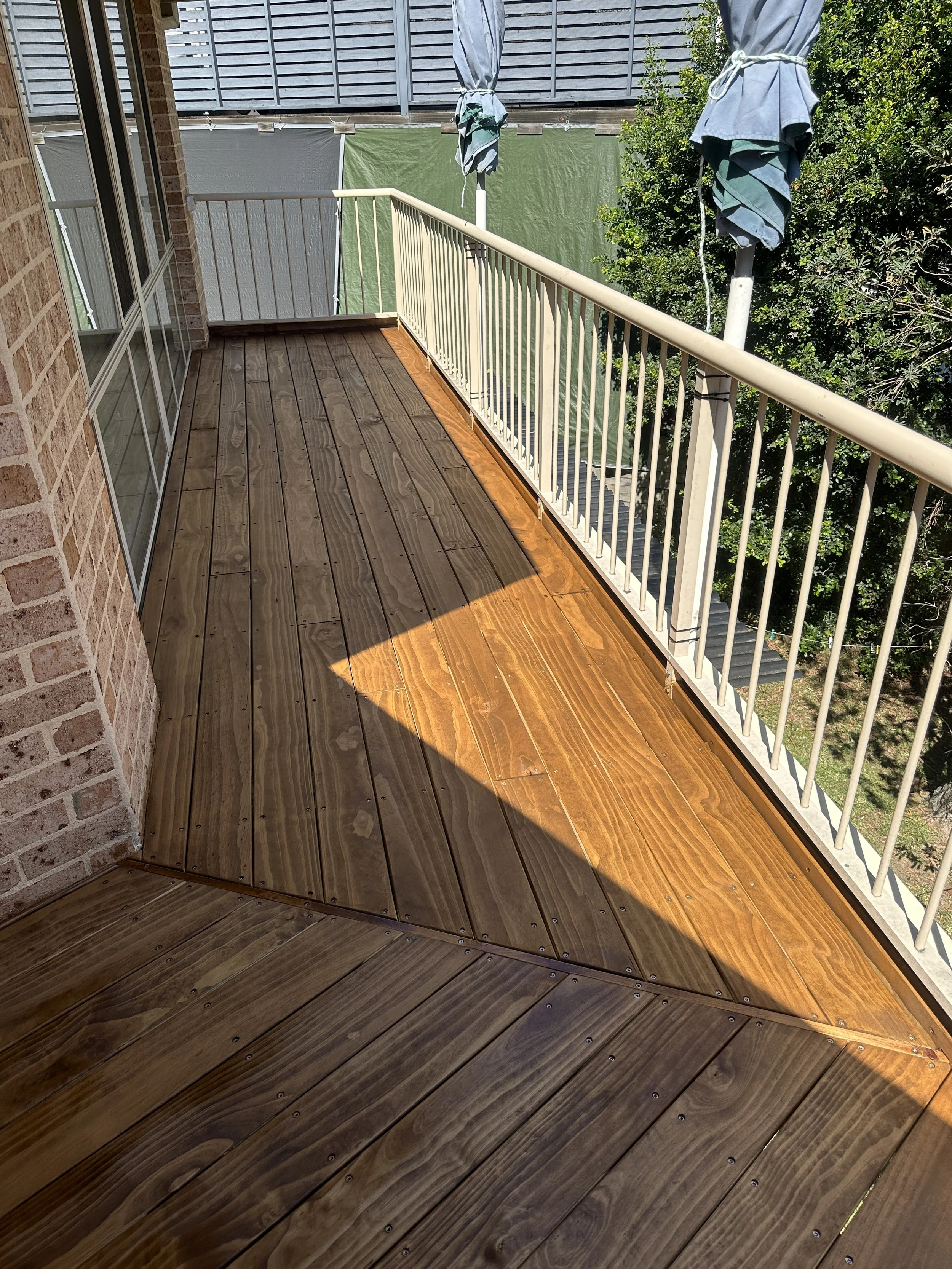 A view from a balcony with wooden flooring, a white metal railing, a brick wall on the left, two blue umbrellas, and green trees in the background.