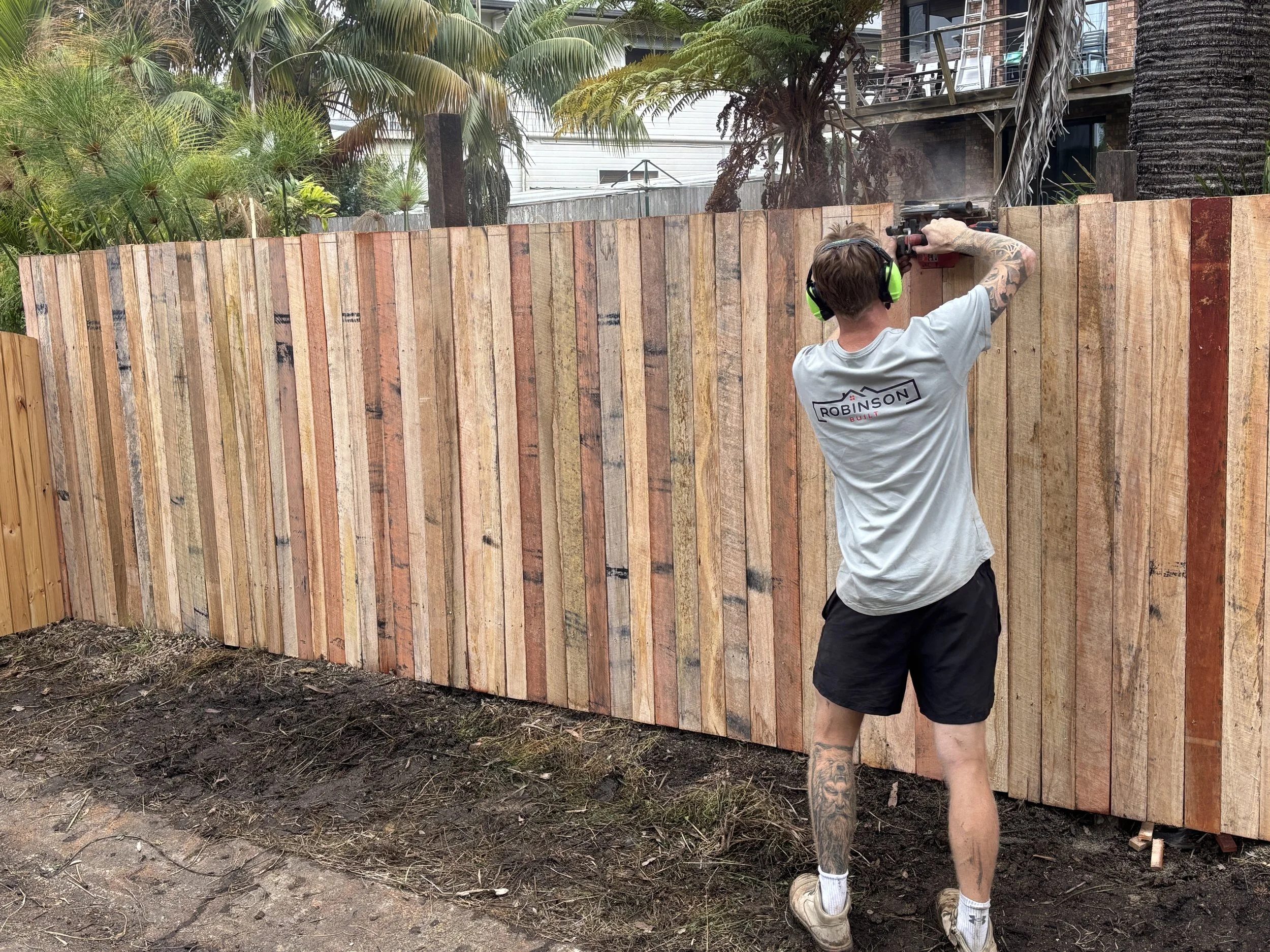A person with tattoos wearing headphones, a gray t-shirt, and shorts, is using a nail gun to attach vertical wooden planks to a curved wooden fence in a backyard with trees and a house in the background.