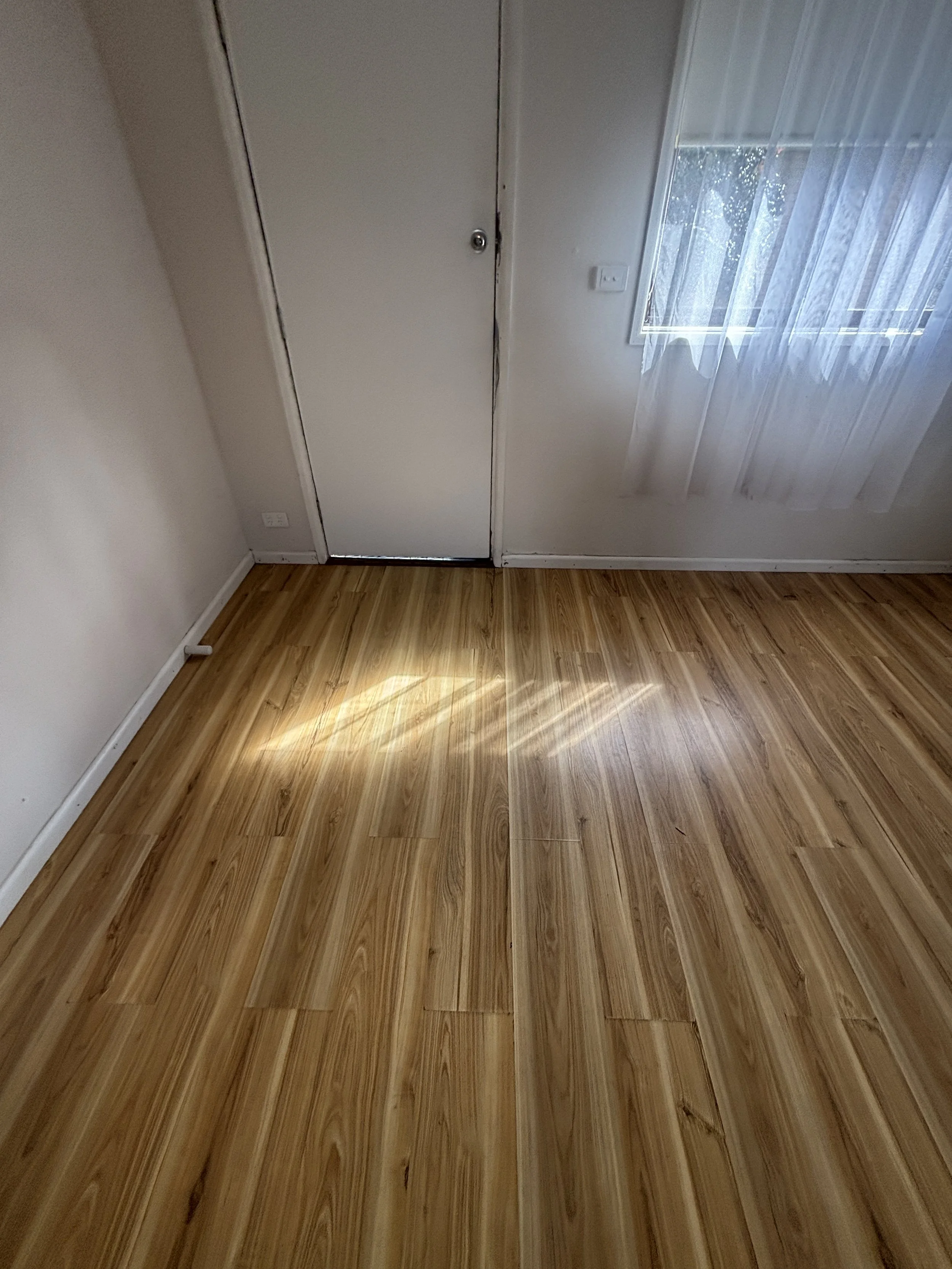 Empty room with wooden flooring, white walls, a door, and a window with sheer curtains, sunlight casting on the floor.