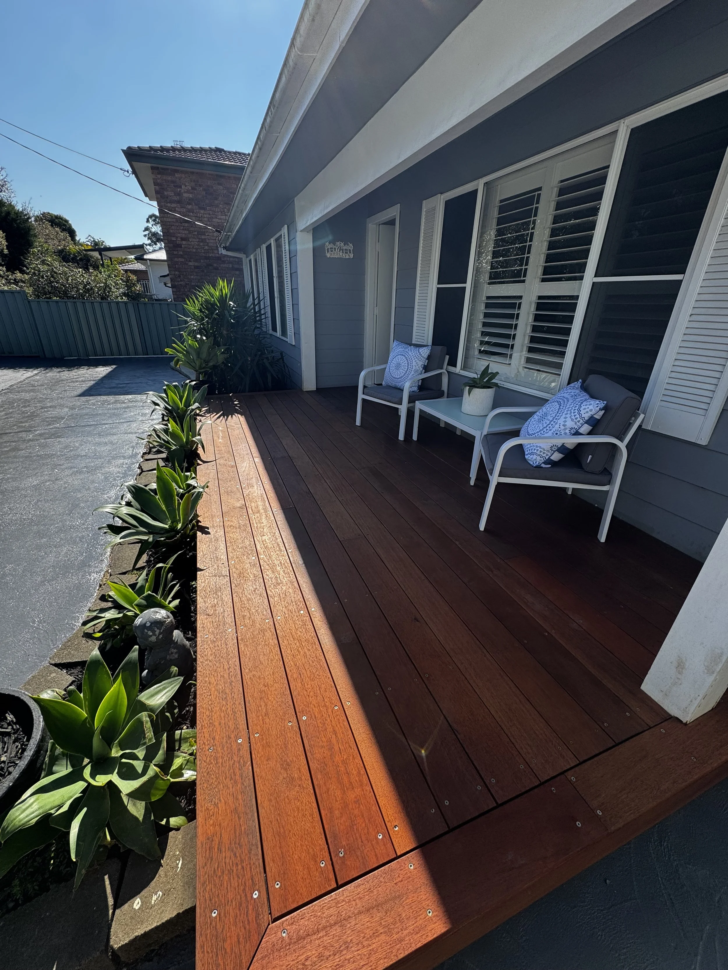 Front porch with two white chairs, pillows, potted plants, and wooden decking next to a house with large windows and shuttered blinds.