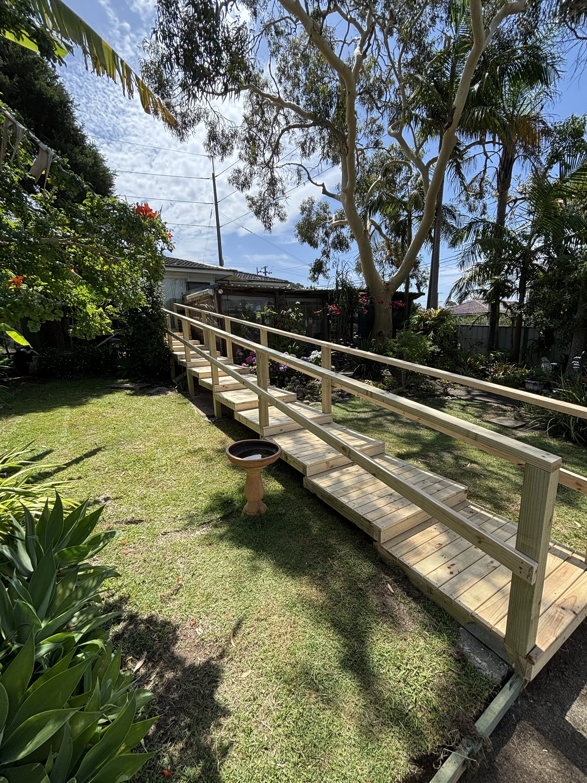 A backyard garden with a partially constructed wooden ramp, surrounded by lush plants and trees, with a house in the background and a bright blue sky with scattered clouds.