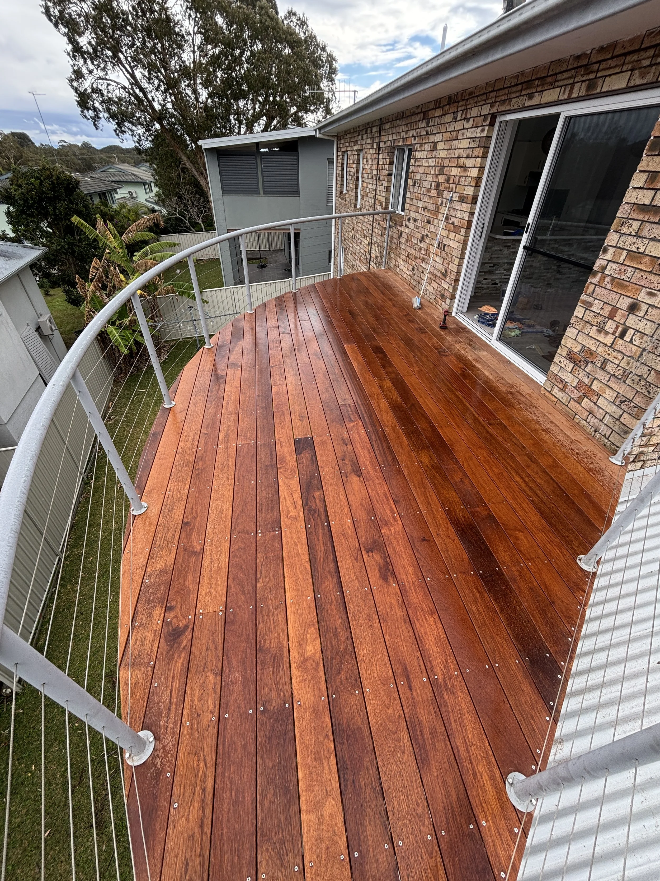 A balcony with polished wooden flooring, a metal railing, and a sliding glass door leading into a brick house. The background shows trees and neighboring houses under a partly cloudy sky.