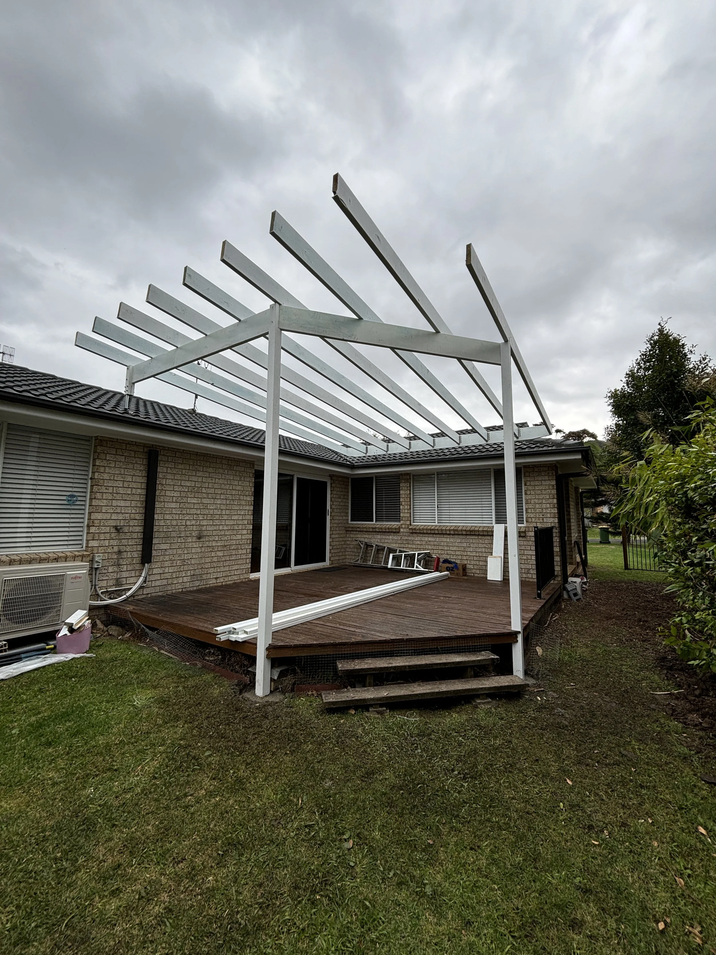 Incomplete white wooden structure being assembled on a backyard deck with a brick house behind, cloudy sky overhead, and some gardening tools and materials on the ground.