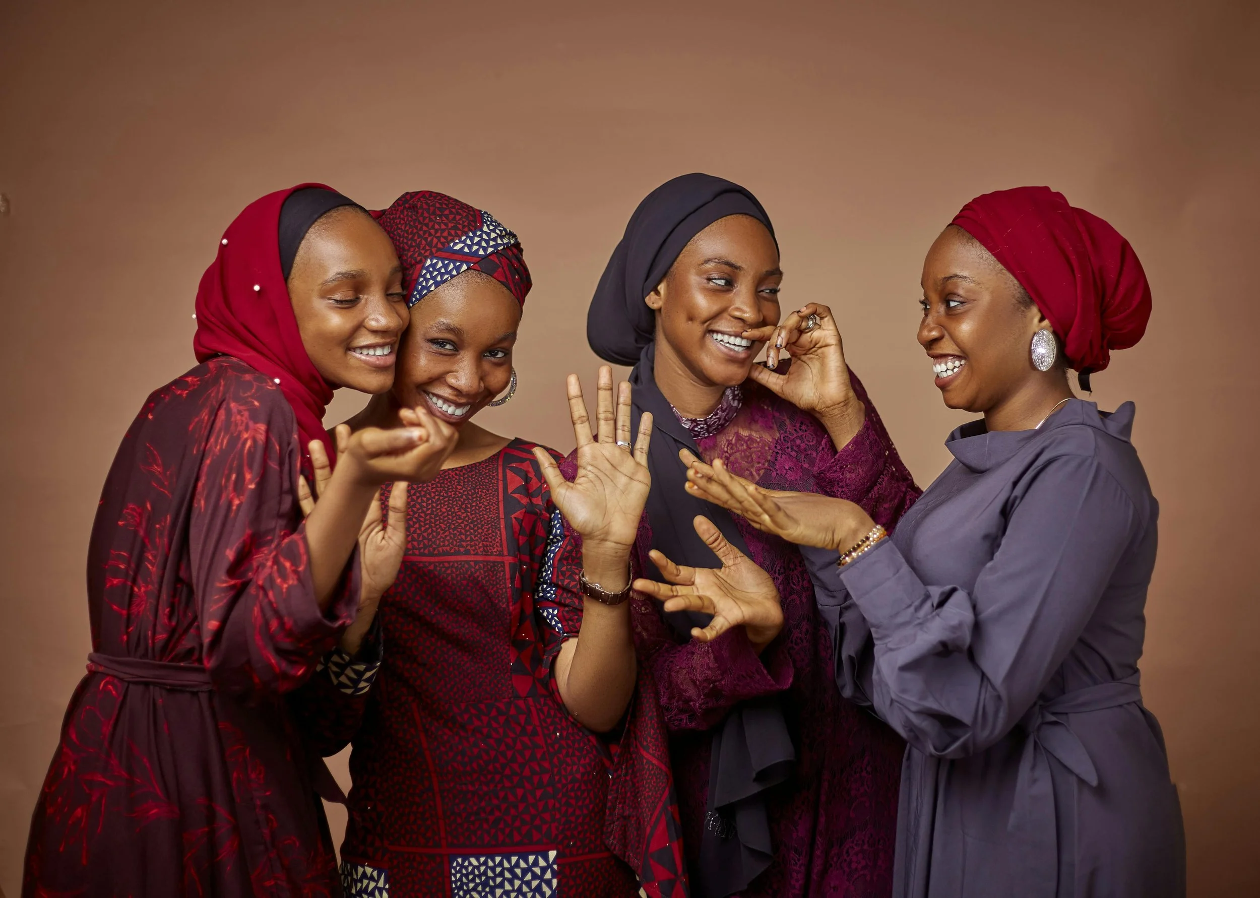 Four women of African descent dressed in colorful traditional attire, smiling and engaging in lively conversation against a neutral background.
