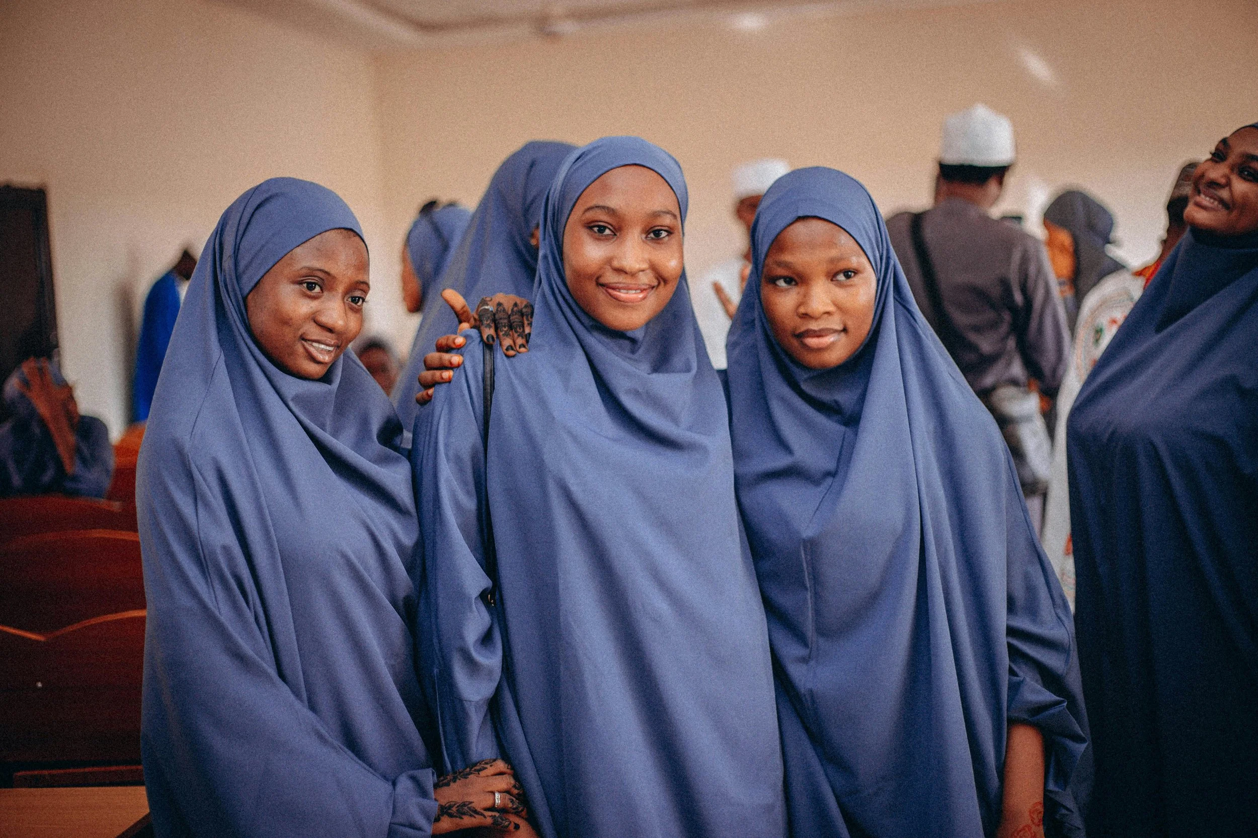 Three women in blue hijabs and robes, smiling, standing close together at an indoor event, with more people in the background.