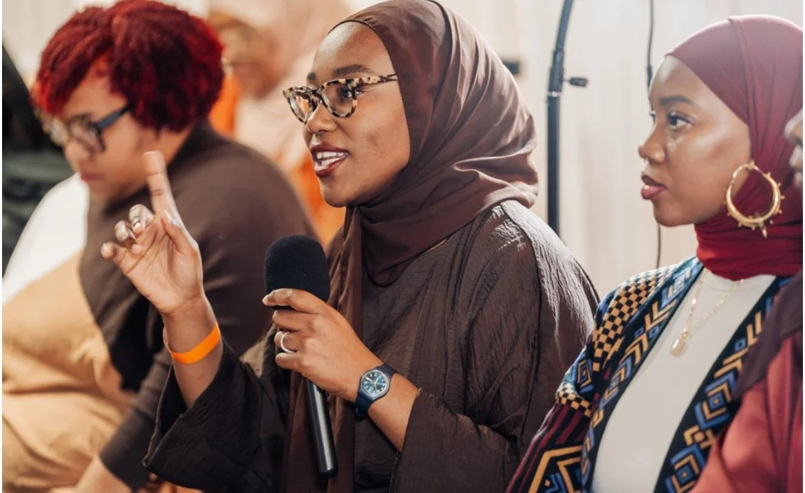 A woman wearing a hijab and glasses holding a microphone and speaking during a discussion or panel.