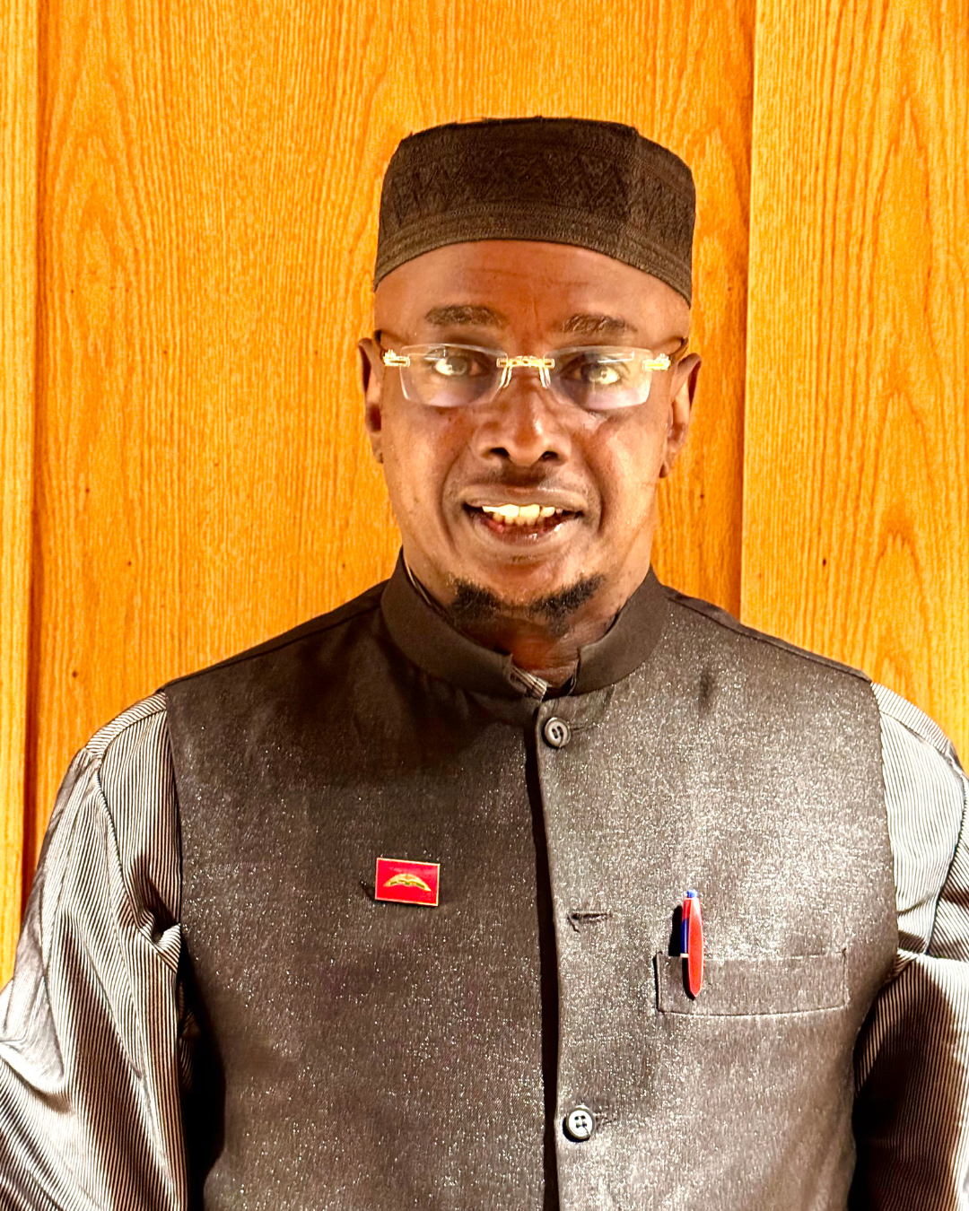 Man in traditional attire wearing glasses, a black hat, and a lapel pin, standing in front of a wooden background.