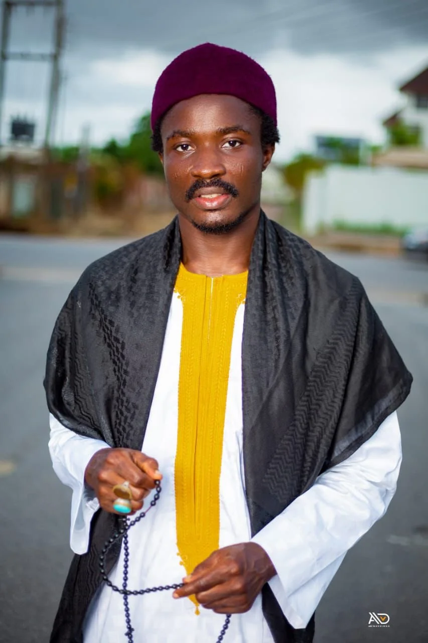 A man wearing a red cap, white and yellow traditional attire, and a black shawl, standing outdoors on a cloudy day.