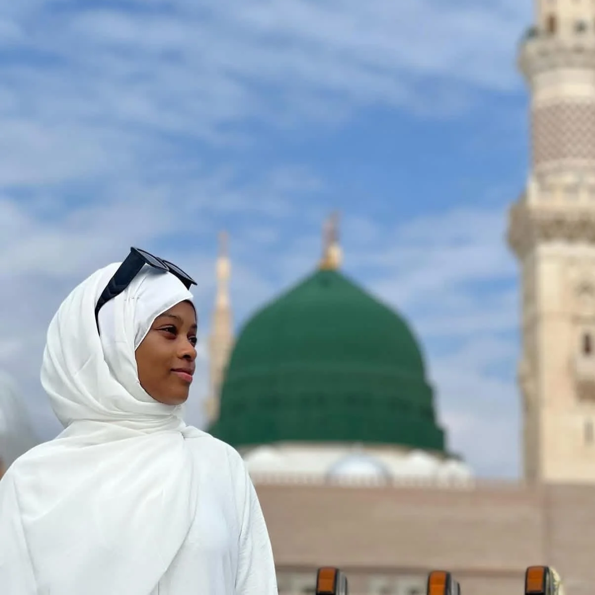 Young woman wearing a white headscarf and sunglasses perched on her head, standing in front of the Al-Masjid an-Nabawi mosque in Medina, Saudi Arabia, with a green dome and minarets in the background.