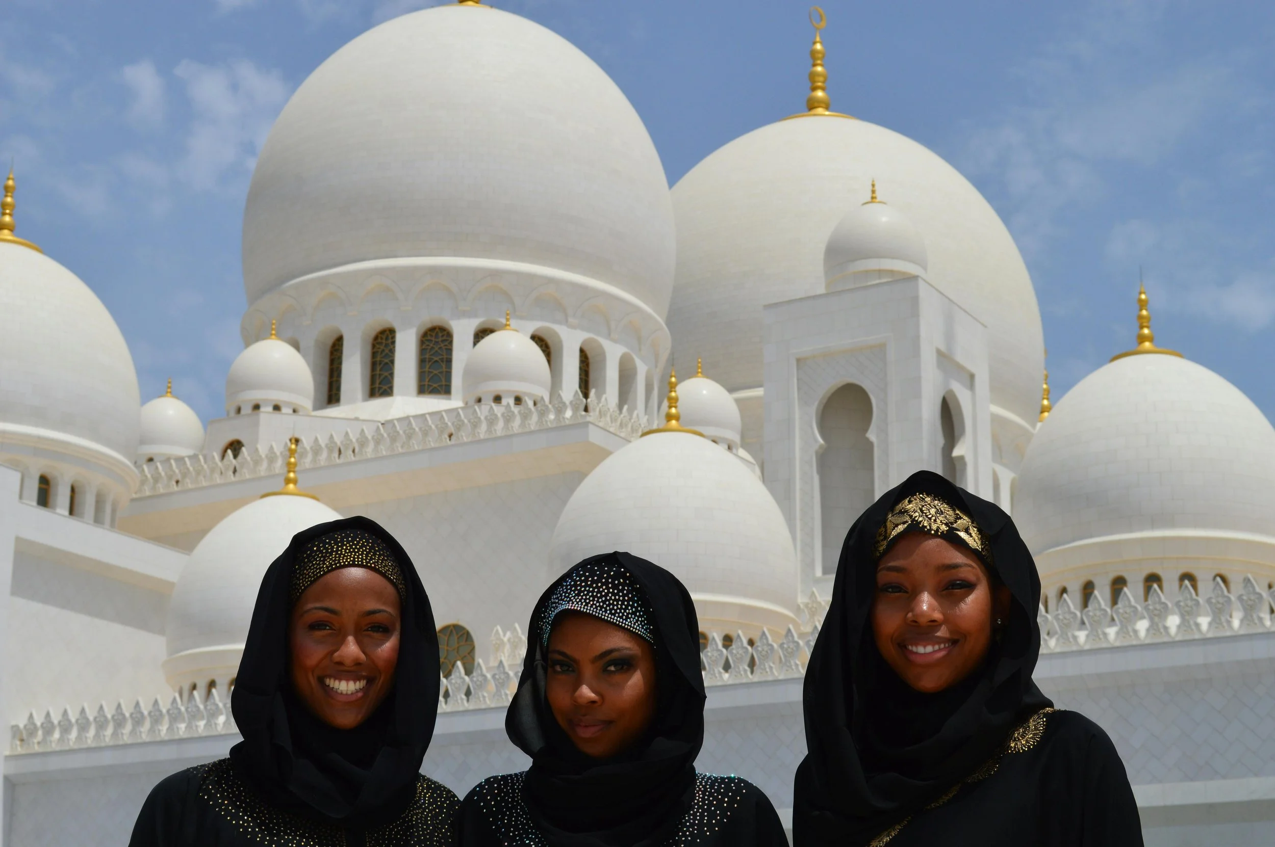 Three women dressed in black traditional attire with head coverings, standing in front of the white domed architecture of the Sheikh Zayed Grand Mosque in Abu Dhabi, United Arab Emirates.