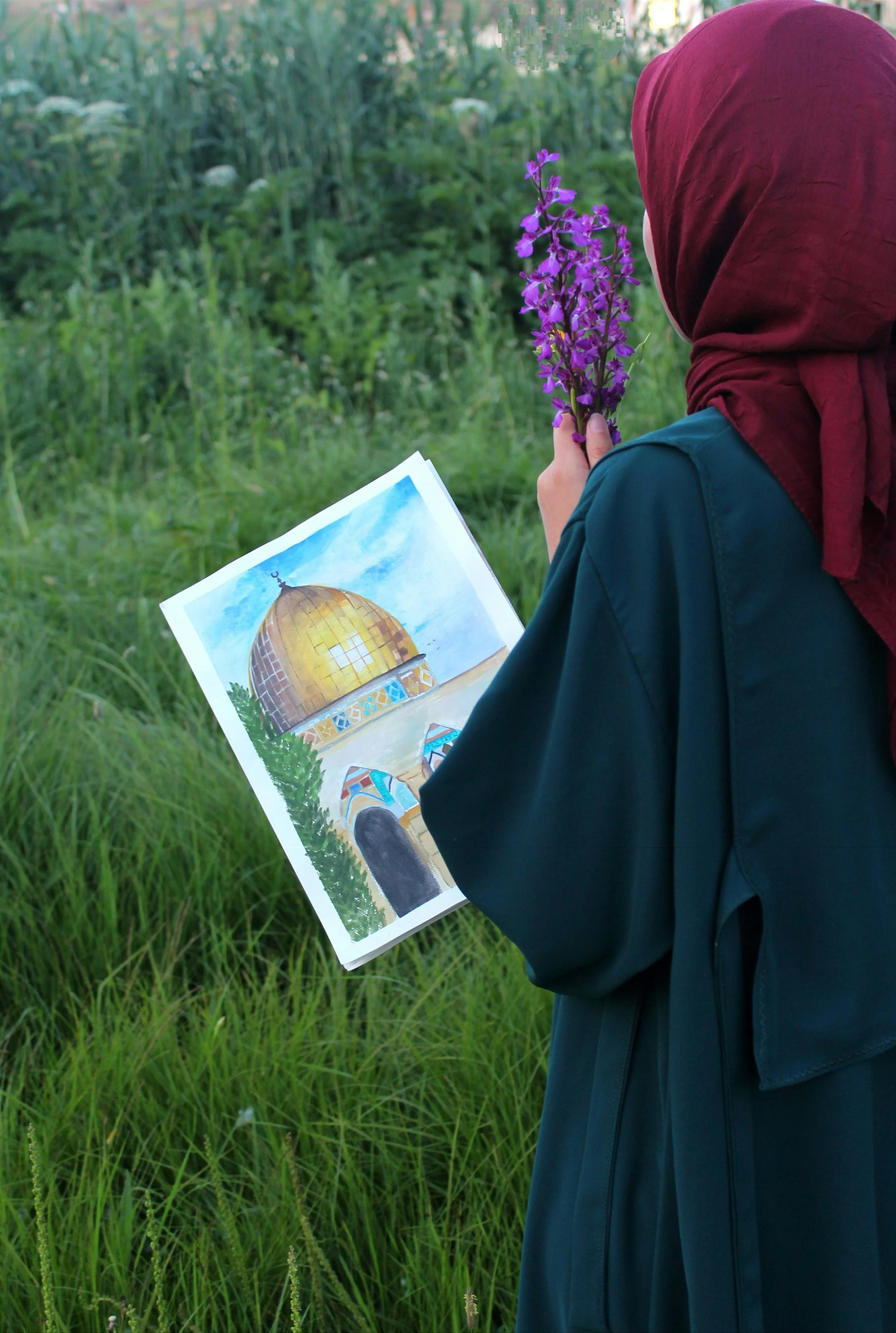 A person wearing a red headscarf and dark green coat is standing in a grassy field, holding a bunch of purple flowers in one hand and a watercolor painting of a golden-domed building in the other.