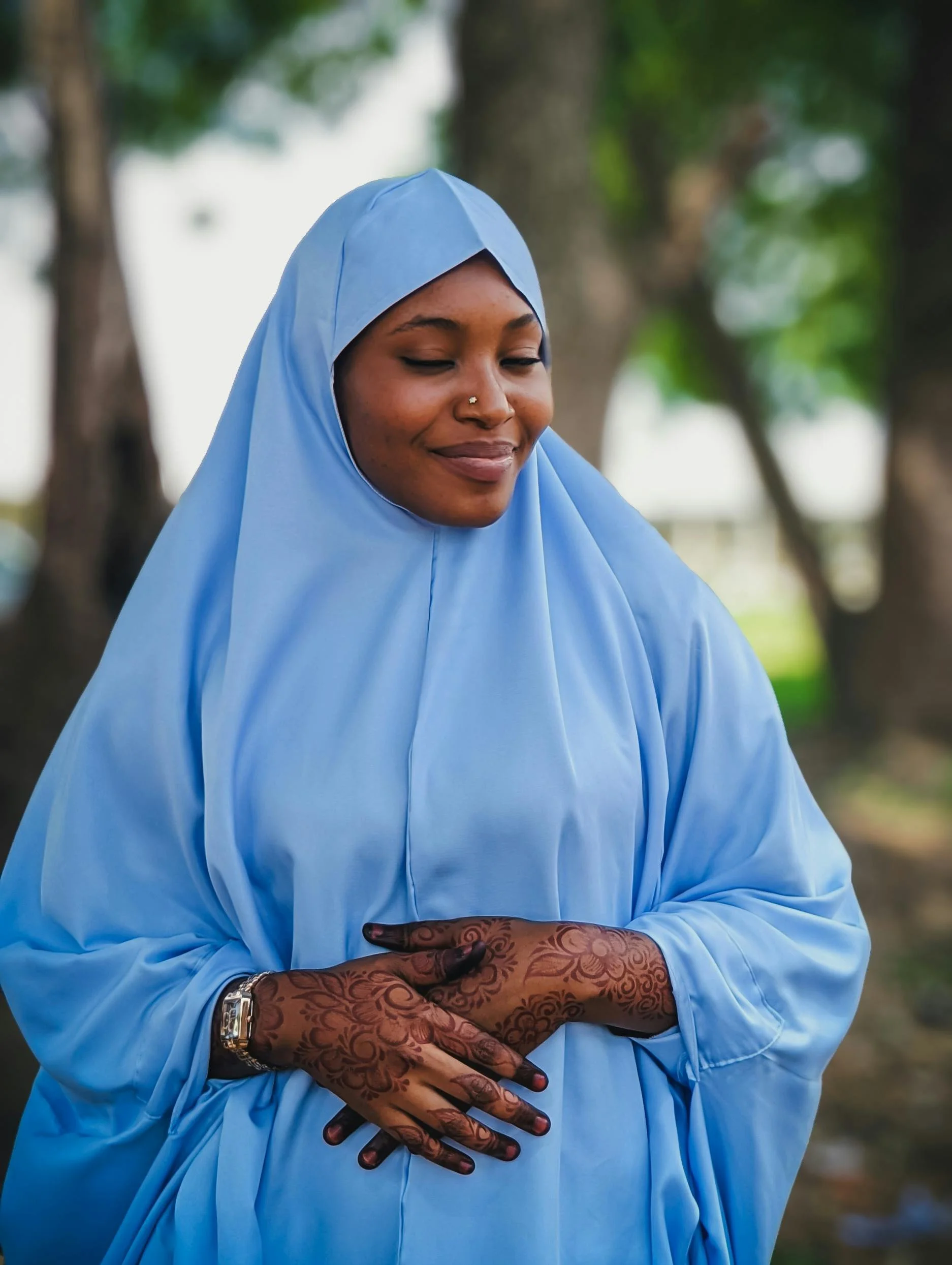 A woman dressed in a blue hijab and matching loose clothing outdoors, smiling with eyes closed, showing Henna designs on her hands.