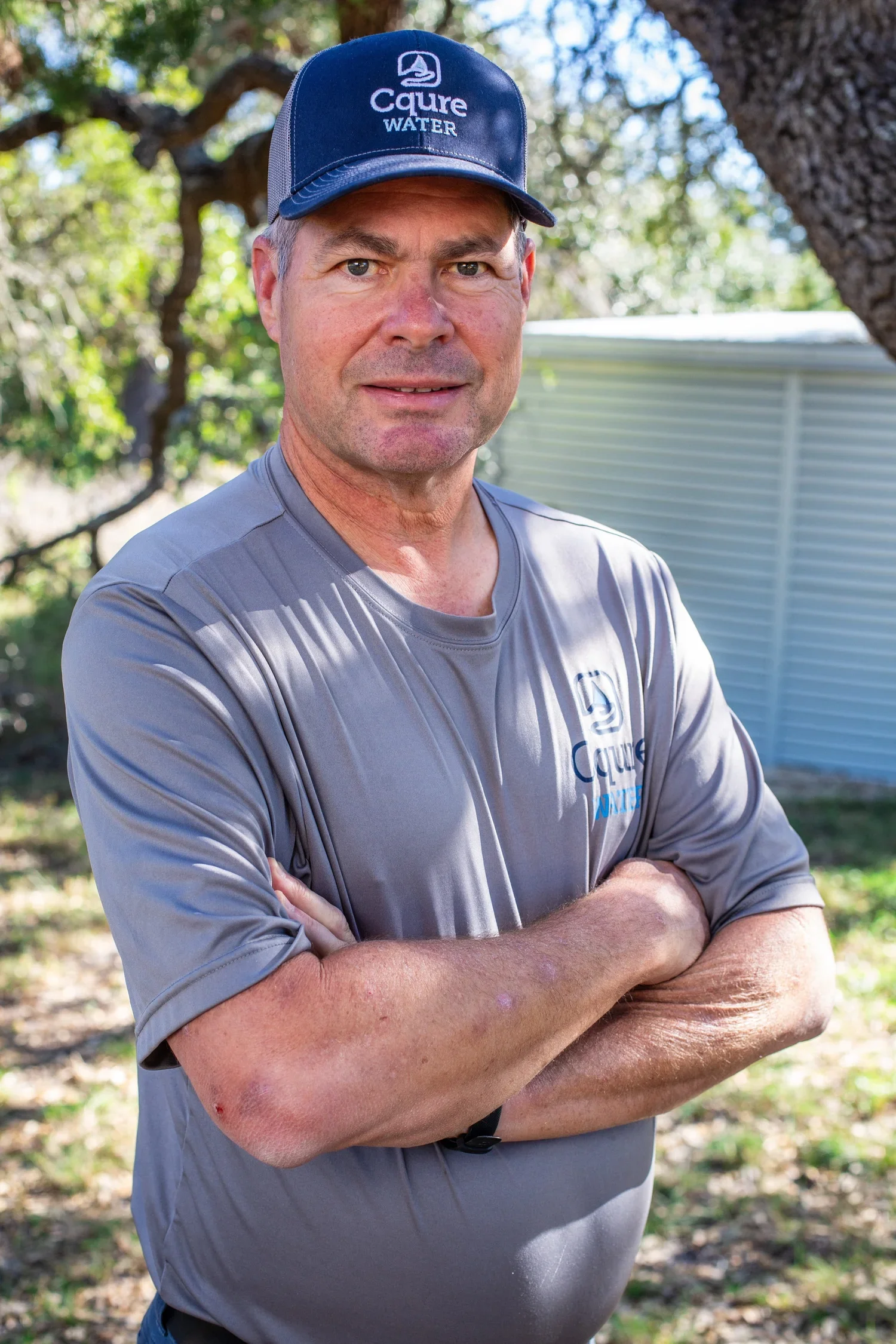 A man wearing a blue cap with the words 'Caire Water' and a gray sports shirt with similar logo, standing outdoors with arms crossed in front of a tree and a light-colored shed.