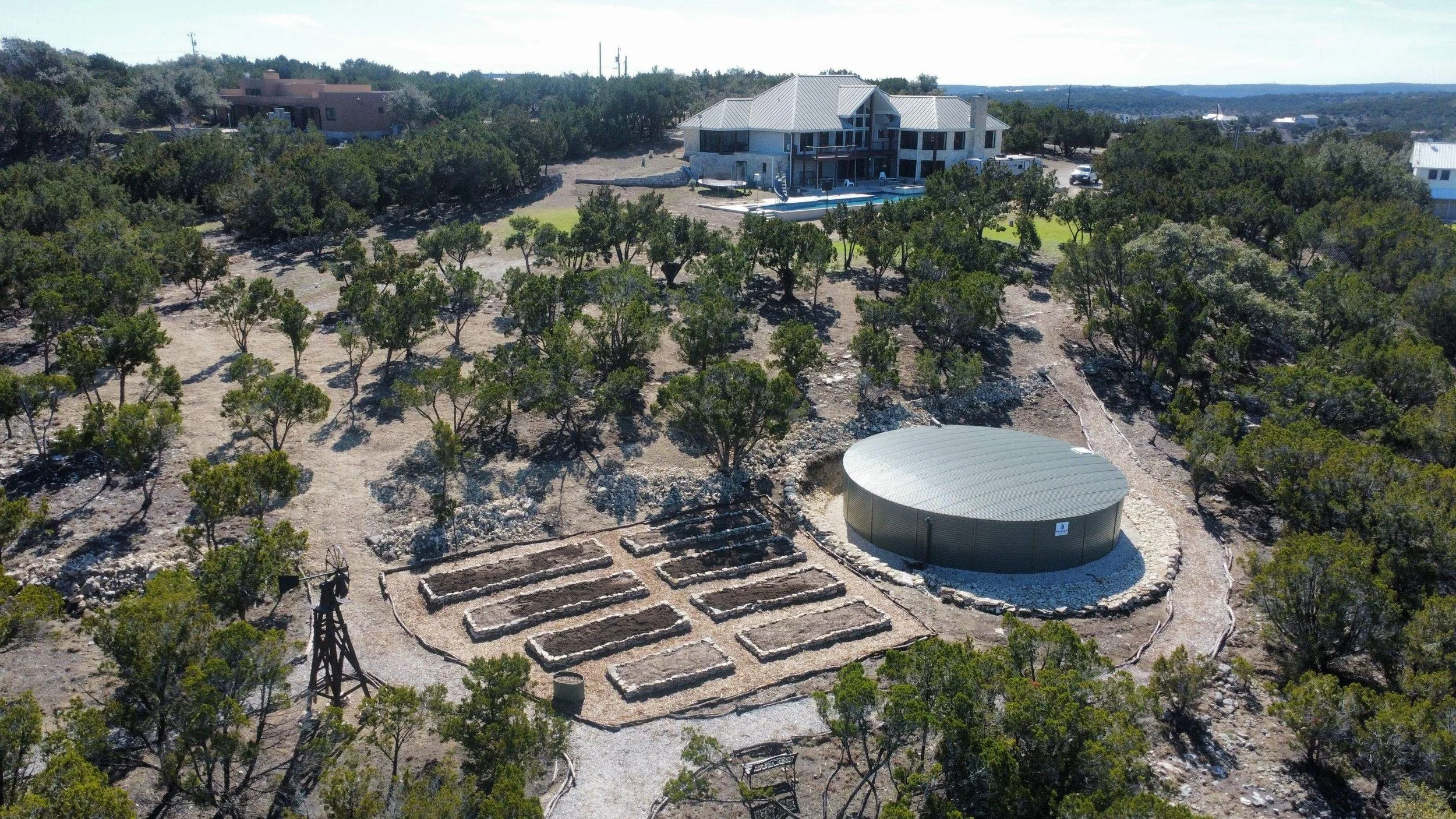 Aerial view of a rural property with a large modern house, a round metal storage tank, small garden plots, and surrounding trees in a dry, hilly landscape.