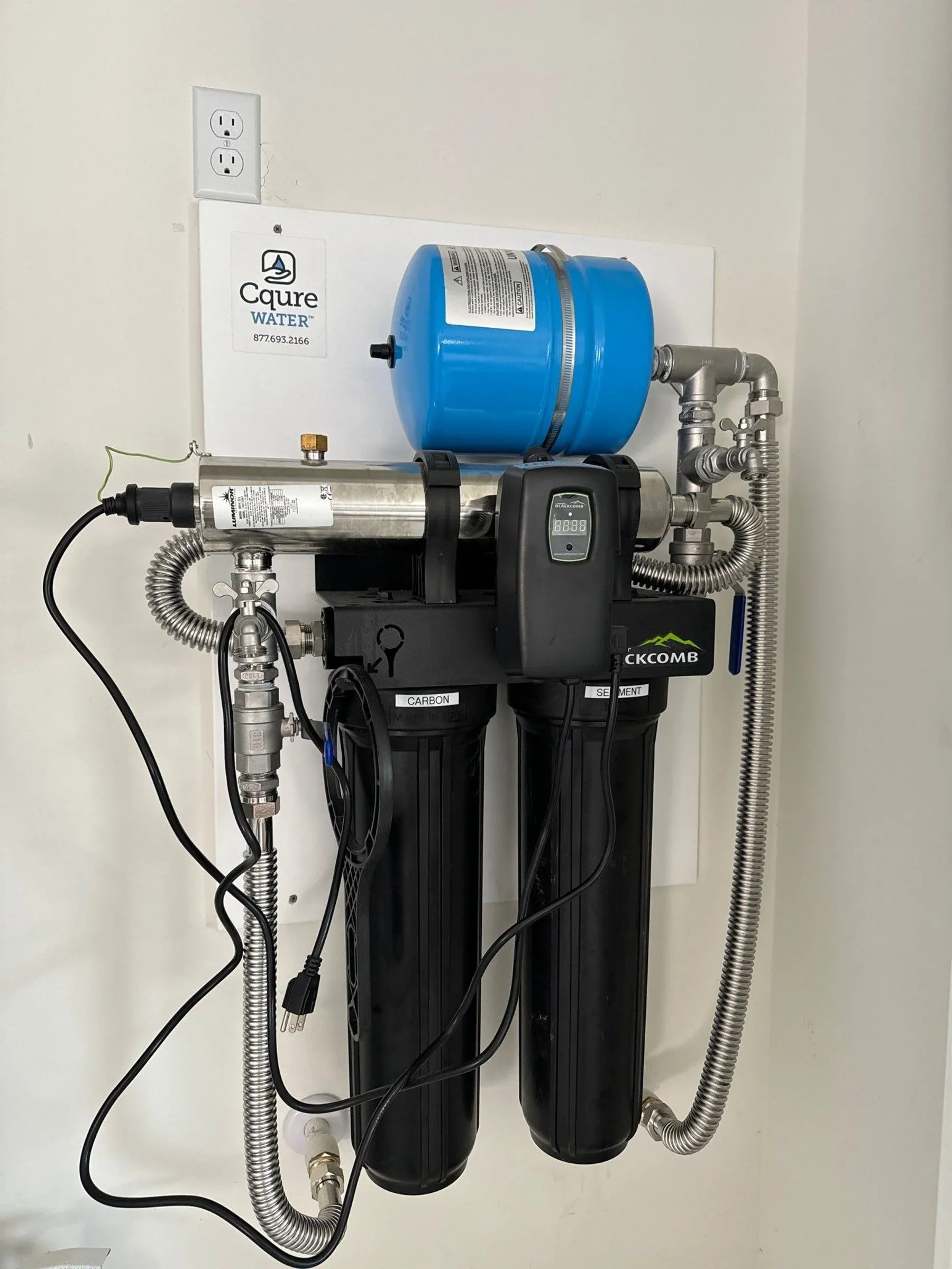 Water filtration system with two black filters labeled 'Carbon' and 'Sediment,' a blue pressure tank, and connected metal and flexible hoses against a plain white wall.