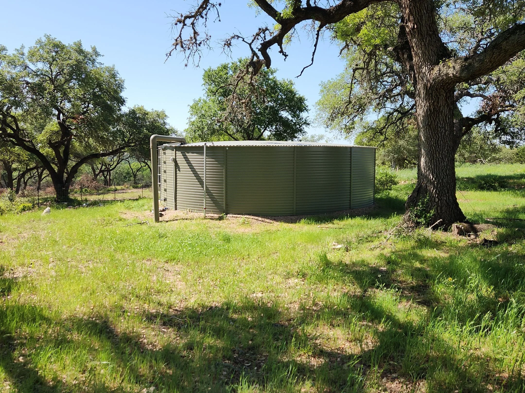 A metal water tank outdoors among trees with lush green grass, under a clear blue sky.