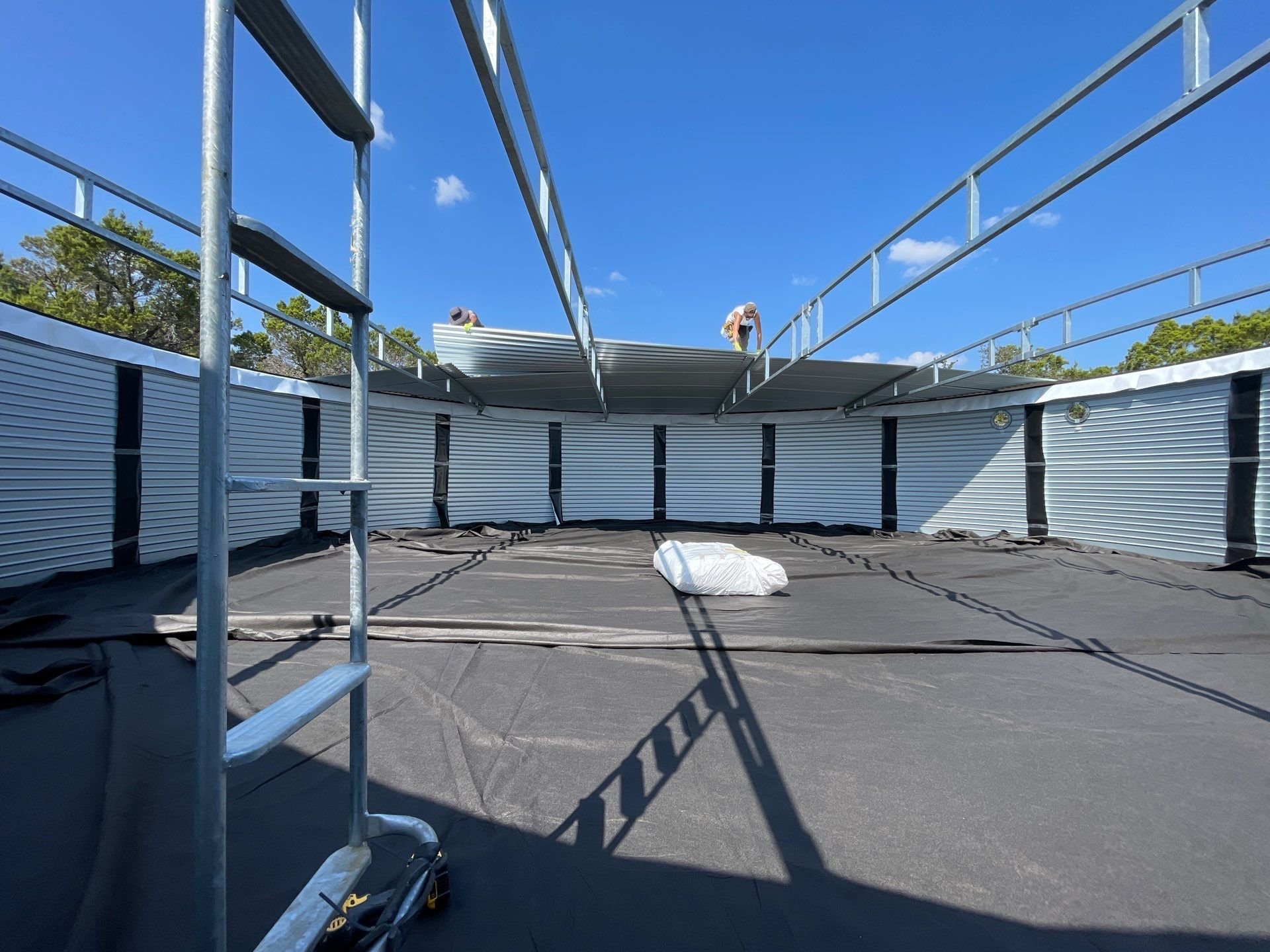 Construction workers install a metal roof on a building with a curved wall. The photo is taken from inside the structure, with scaffolding and construction tools visible on the ground. The sky is clear and blue.
