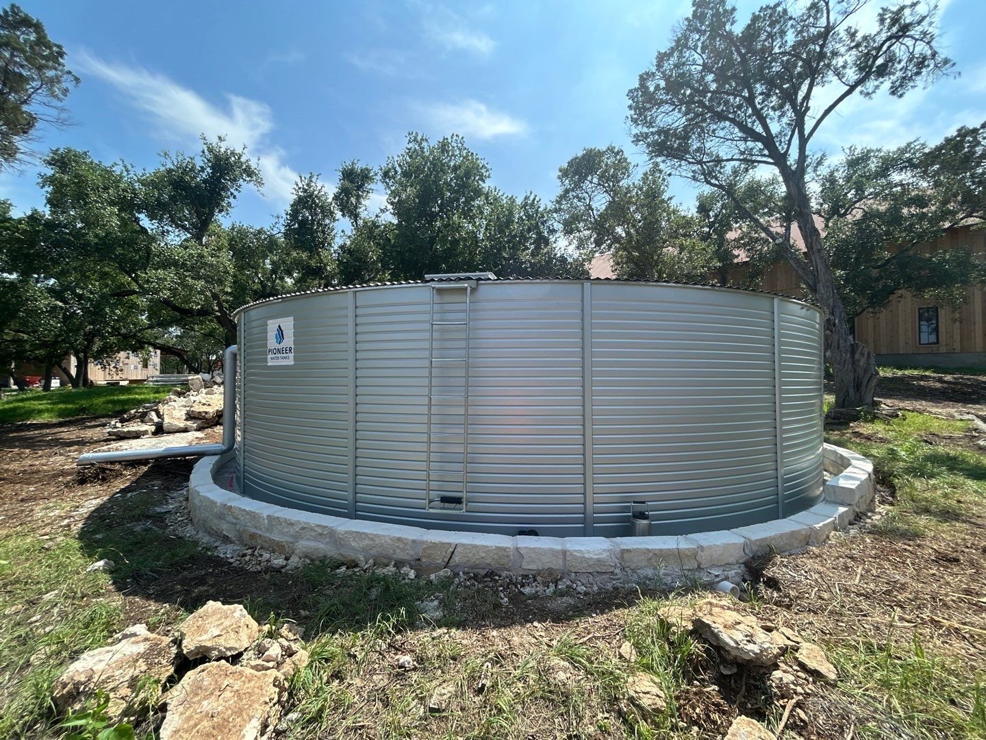 A large, round water storage tank made of metal, situated outdoors on a concrete and stone foundation, with trees and a building in the background under a partly cloudy sky.