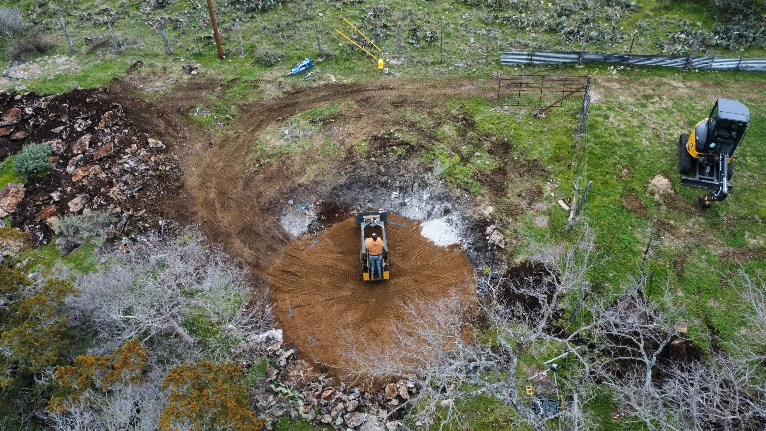 An aerial view of a construction site with a small excavator leveling soil in a circular pattern, a second excavator nearby, and a person operating the equipment. There is a fence and some trees around the site.