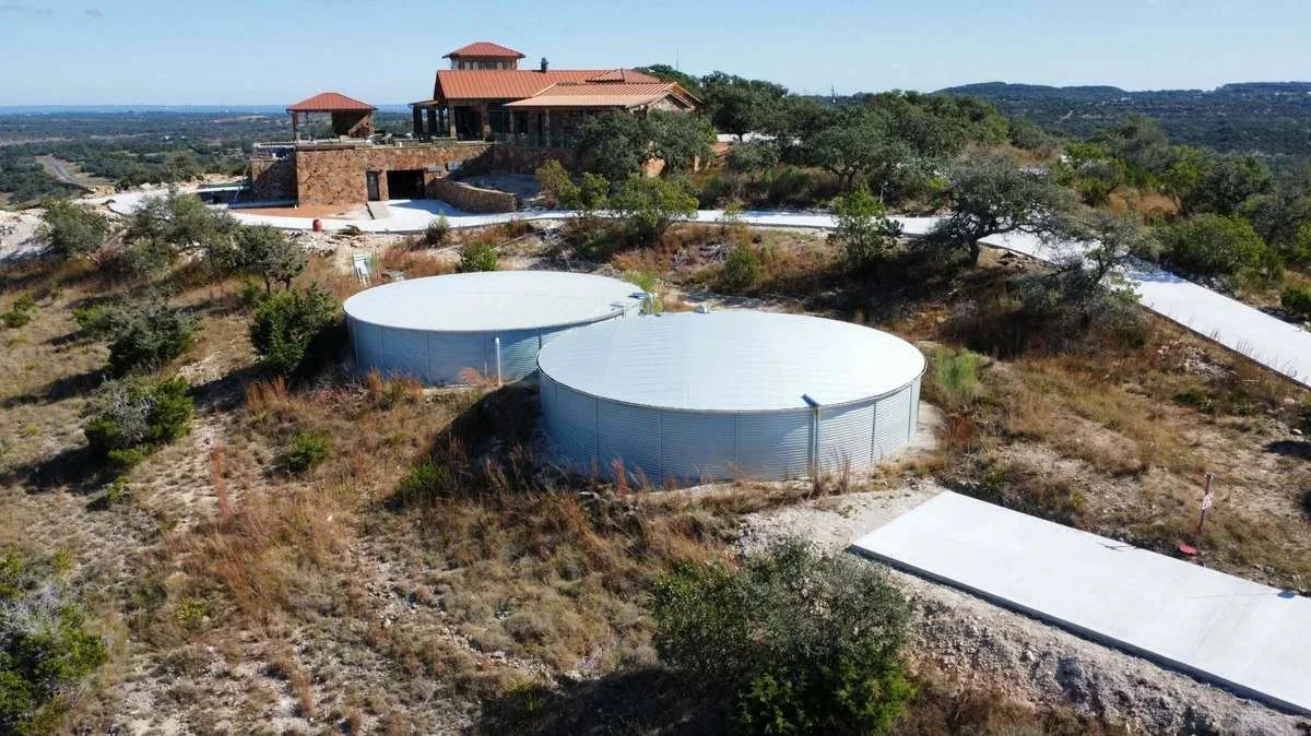 An aerial view of a large house on a hill with a reddish roof, surrounded by trees and bushes, and two large white circular water tanks nearby, with a winding driveway leading to the house.