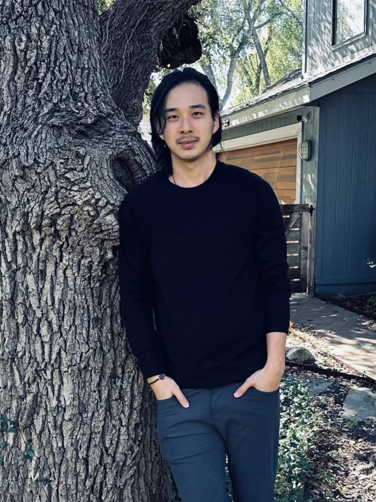 A young man with long black hair and light facial hair standing outdoors next to a large tree with a textured bark. He is wearing a black long-sleeved shirt, dark gray pants, and a watch. The background includes green trees, a blue house, and a partly cloudy sky.