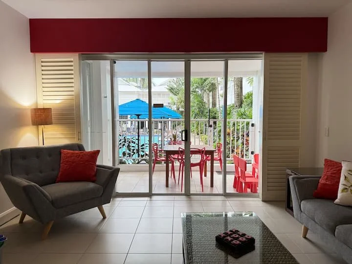 Living room with sliding glass door leading to patio with red and blue furniture and umbrella, overlooking a lush tropical backyard.