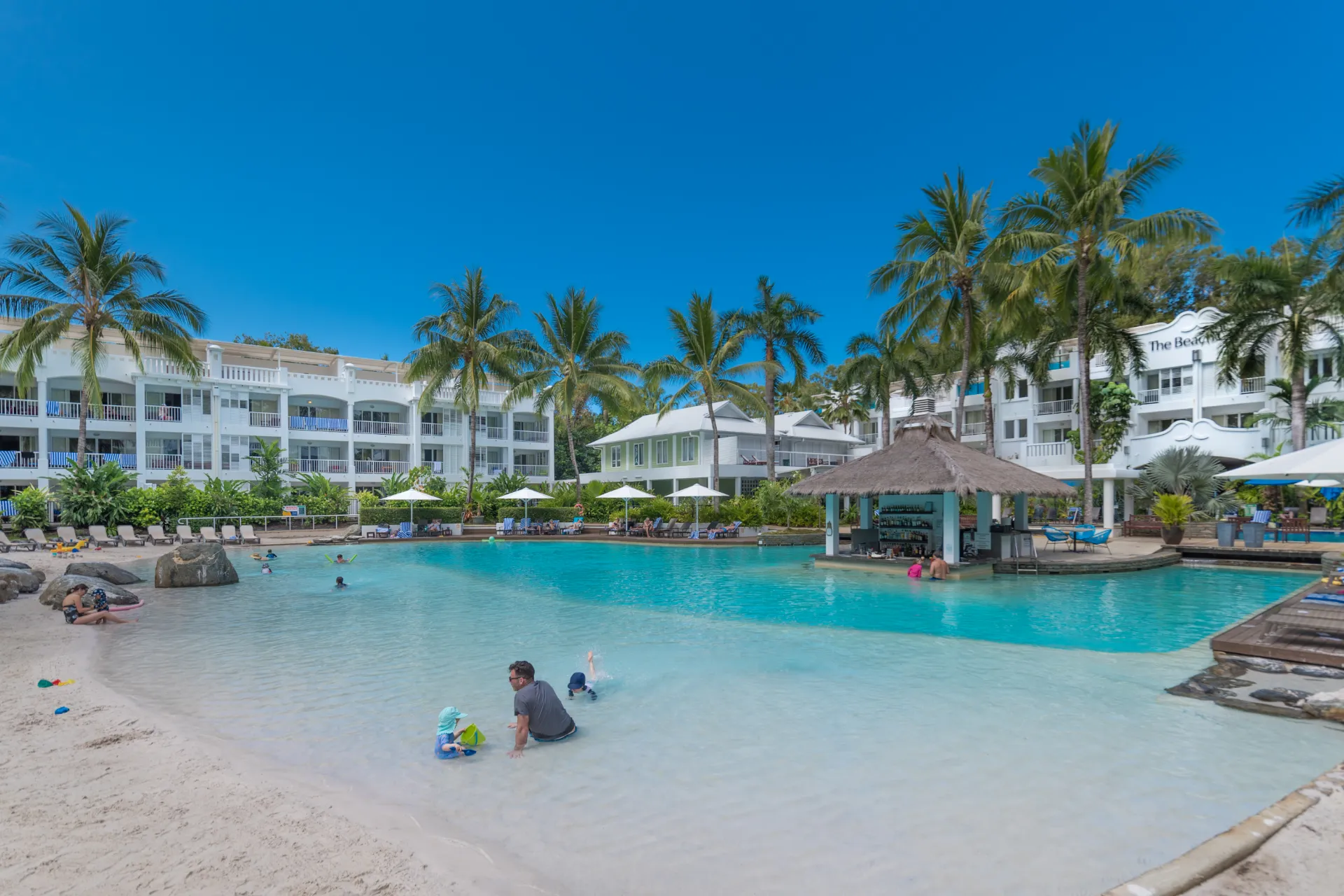 A resort with a large swimming pool surrounded by palm trees, lounge chairs, and white buildings, with people swimming and relaxing.