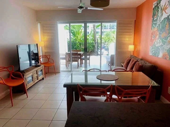 Living room with tiled floor, a TV on a wooden stand, orange chairs, a glass dining table, a brown couch with cushions, and a sliding glass door leading to a balcony with tropical plants outside.
