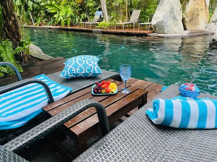 Poolside setup with lounge chairs, towels, and pillows on wooden and wicker surfaces, a glass of water, and a tray of mixed fruit, surrounded by lush green tropical plants.