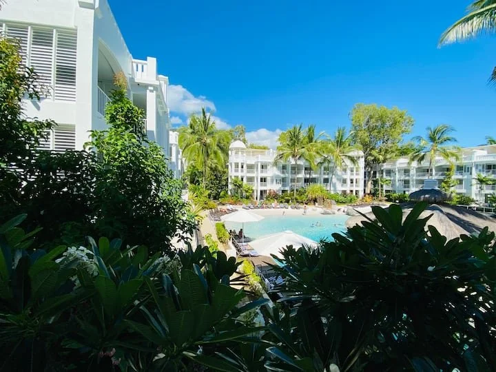 A sunny day at a tropical resort with a large swimming pool, surrounded by white apartment buildings, green palm trees, and lush foliage.