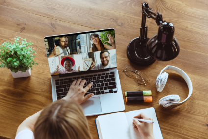 Person participating in a video call on a laptop with six people displayed on the screen, sitting at a wooden desk with a potted plant, desk lamp, headphones, and notebooks.