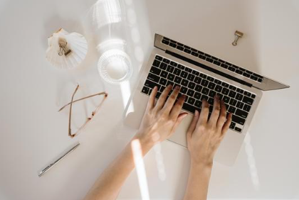 Person typing on a laptop with a white desk, glasses, pen, and a clear drink.