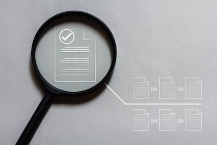 Magnifying glass focusing on a document with a checkmark, with files arranged in a sequence next to it.
