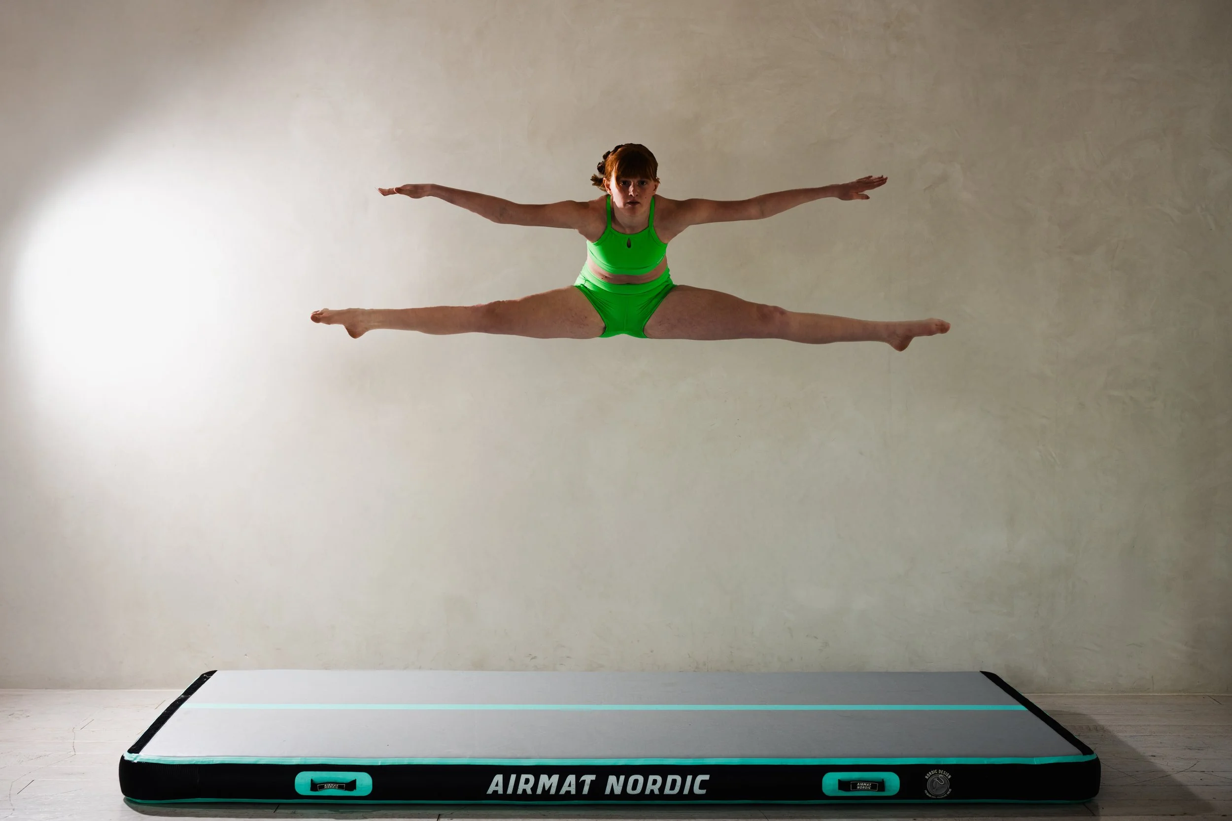 A woman in green athletic wear performing a gymnastic leap in front of a plain wall.