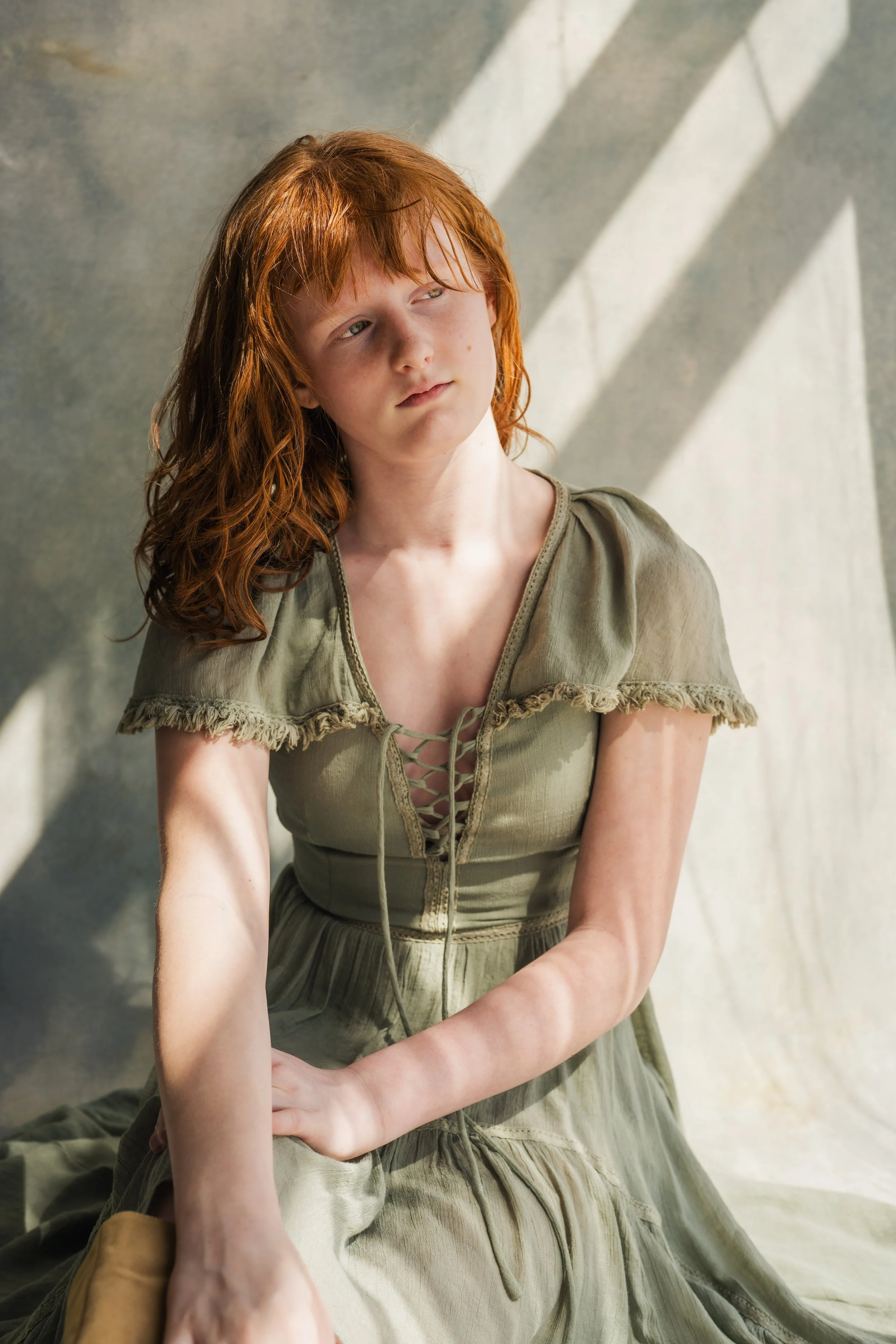 A young girl with red hair and freckles wearing a light khaki dress sitting indoors with sunlight streaming through blinds, casting shadows on the wall behind her.
