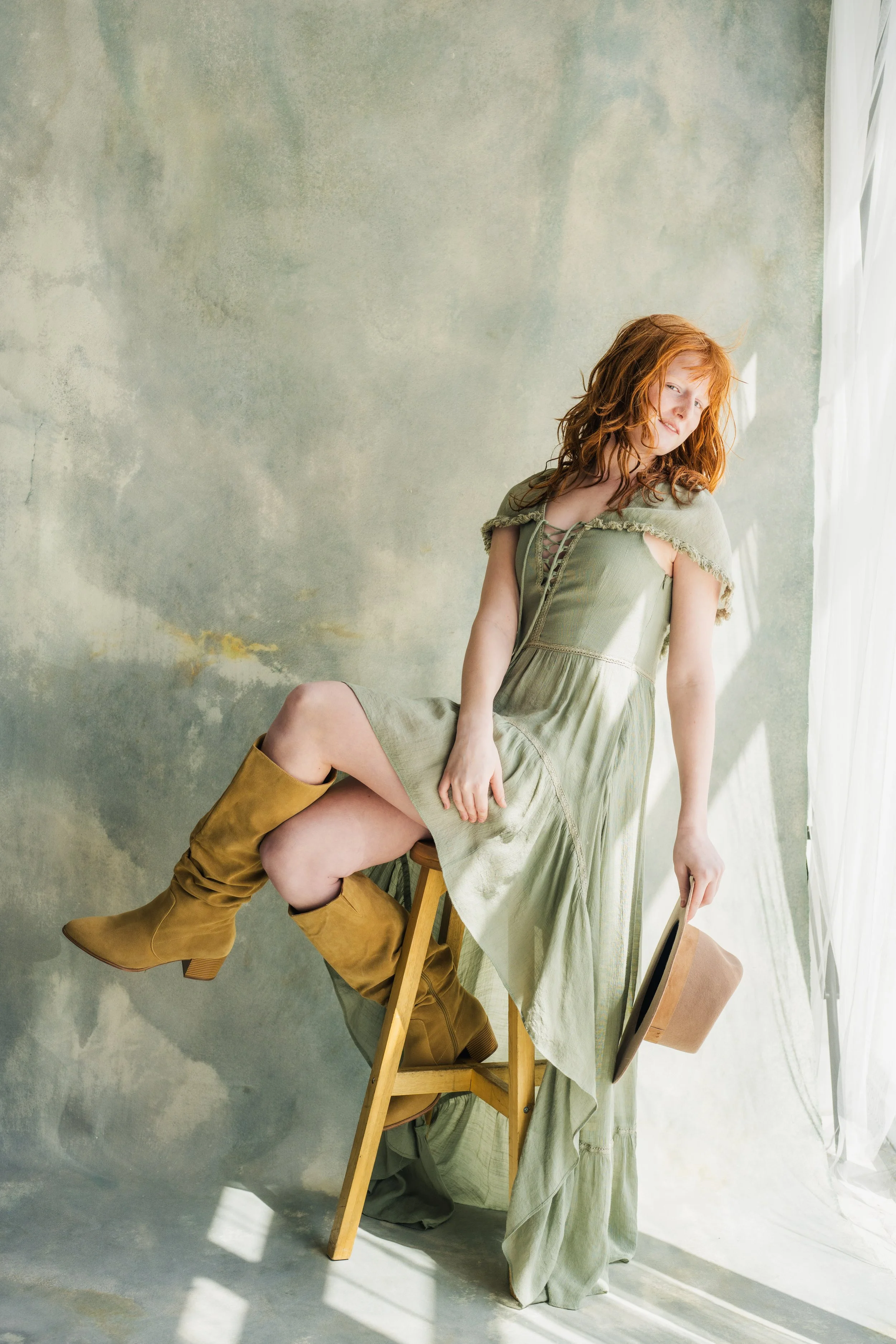 A woman with red hair sitting on a wooden chair, wearing a light green dress and brown knee-high boots, holding a book, in front of a textured, cloudy-green background with sunlight streaming through sheer curtains.