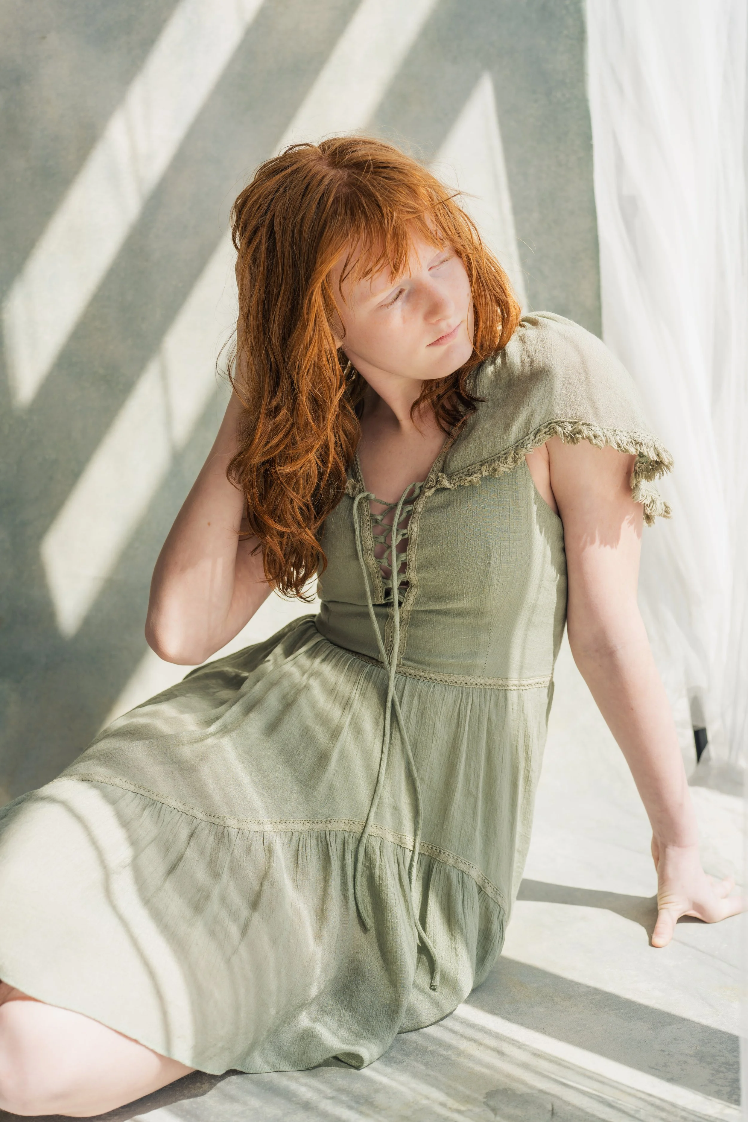 A woman with red hair sitting on the ground, leaning back with one hand inside her hair, wearing a light green dress with lace-up front details. She is posed against a wall with striped shadows.