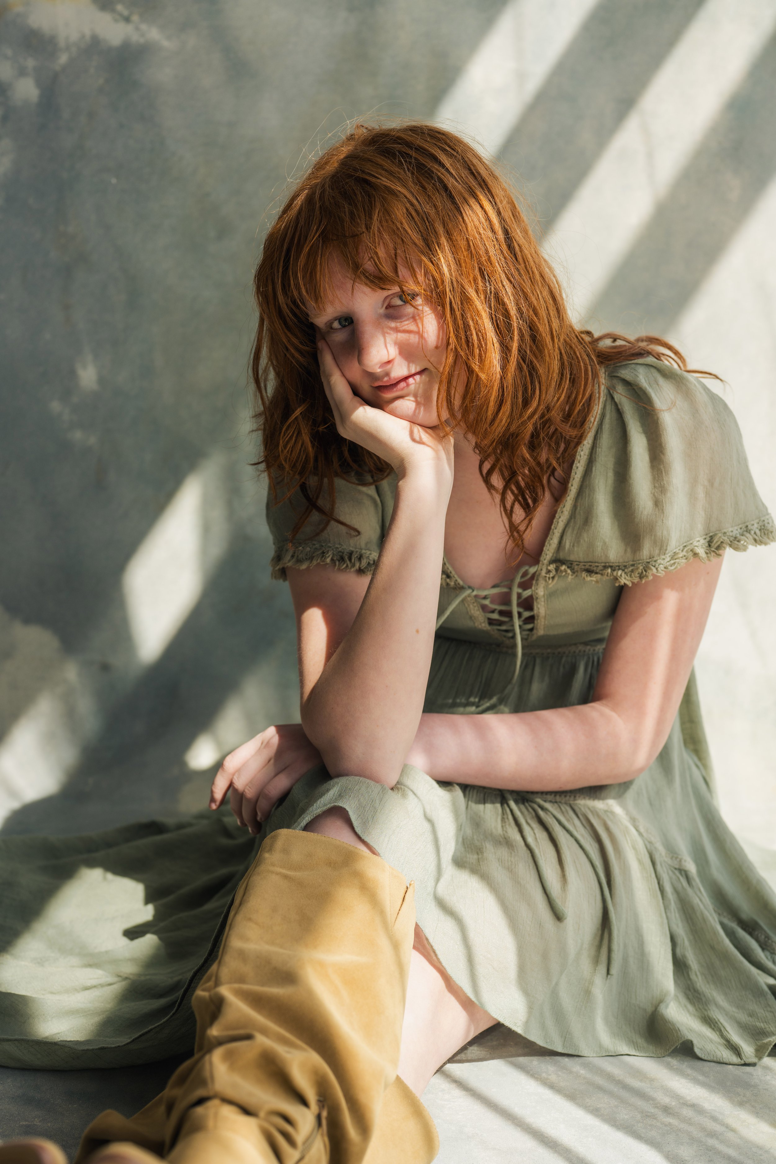 A red-haired woman is sitting on the floor, resting her head on her hand, and looking at the camera with a gentle smile. She is wearing a light green dress and tan boots. The background features a gray wall with sunlight casting shadows.