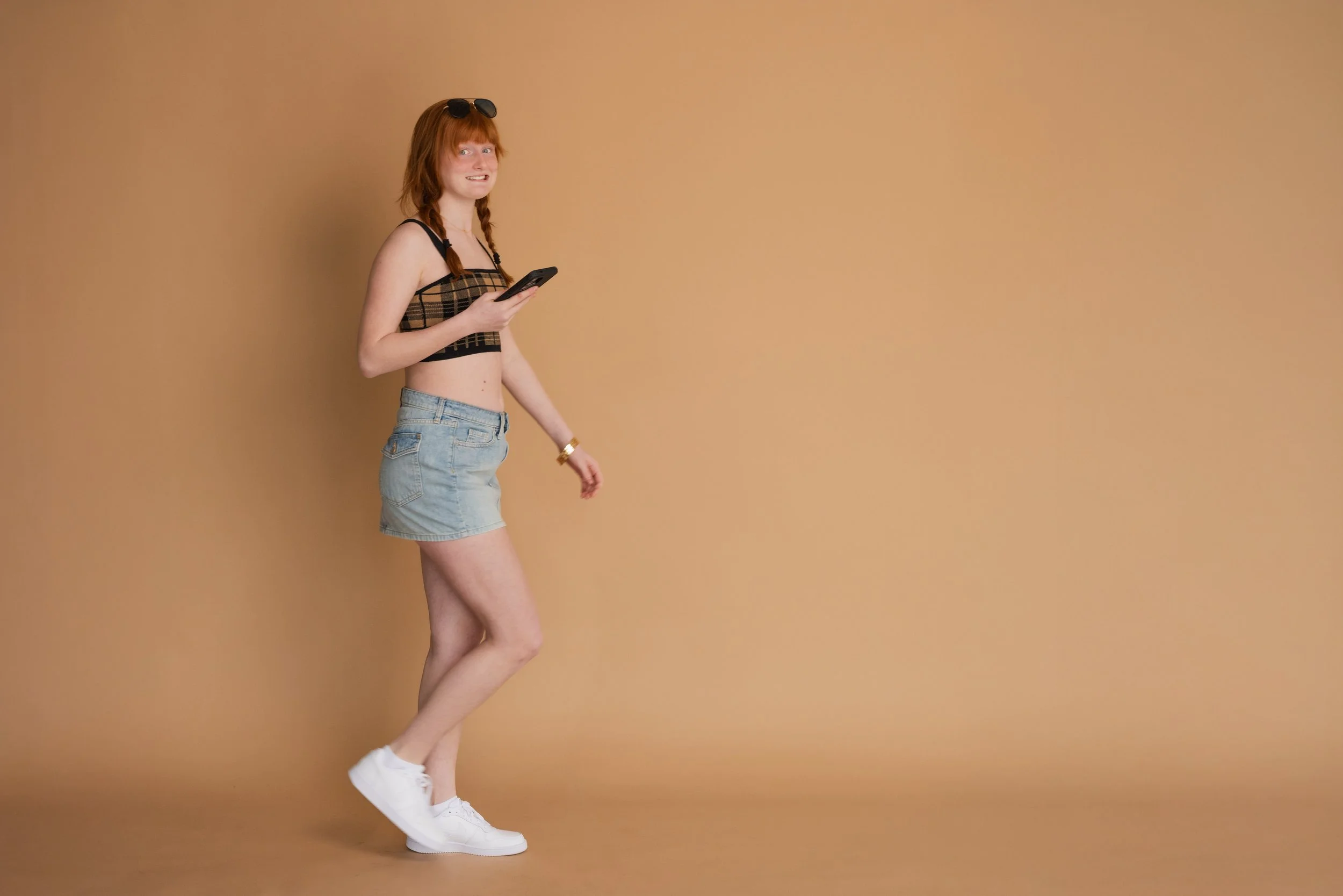 A young woman with red hair, wearing sunglasses on her head, a checkered crop top, denim shorts, and white sneakers, standing against a plain beige background.