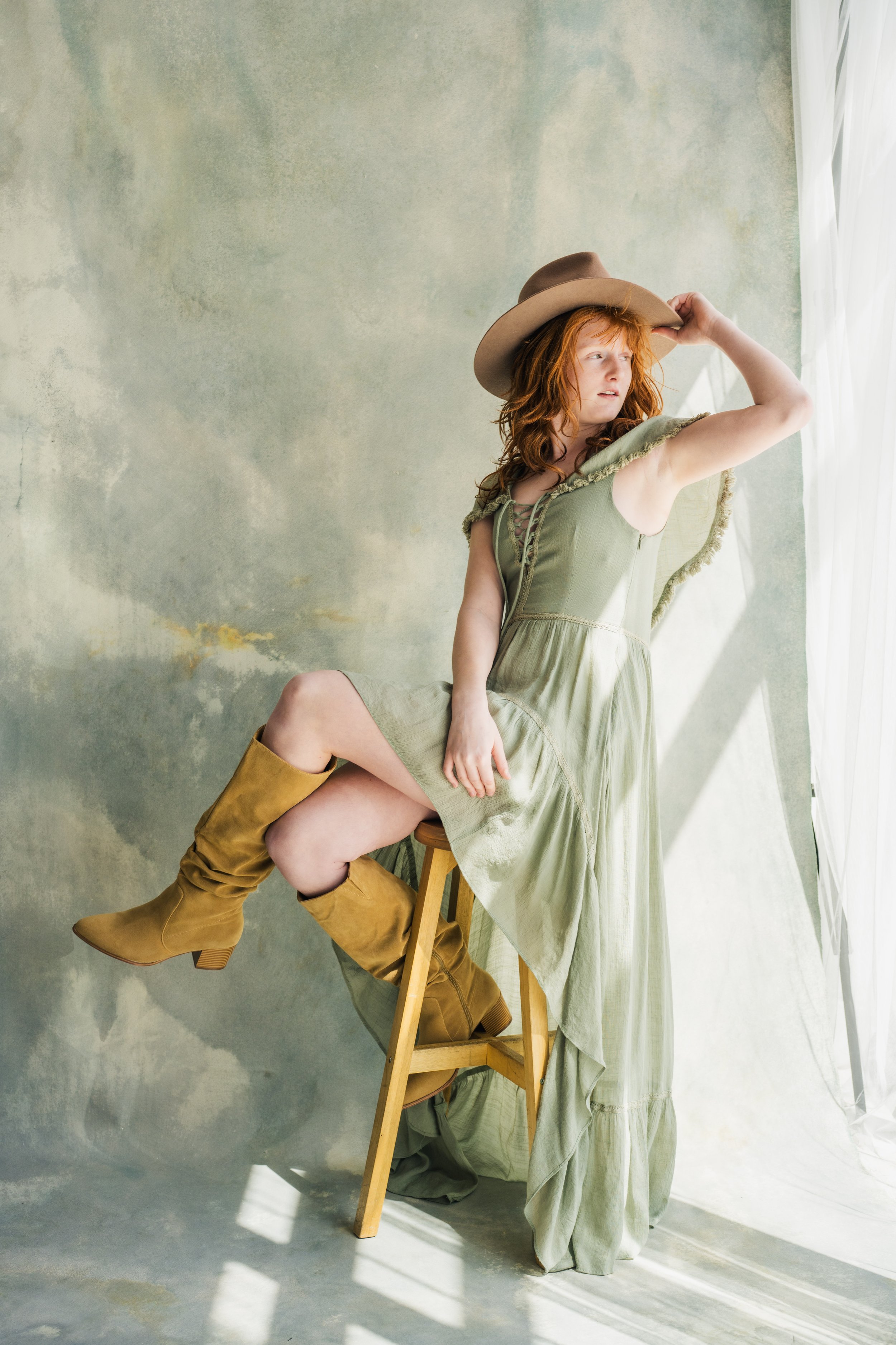 A red-haired woman wearing a long green dress, tan cowboy boots, and a beige wide-brim hat, sitting on a wooden chair in front of a pale green textured wall with white curtains to her right, looking away with her hand on her hat.
