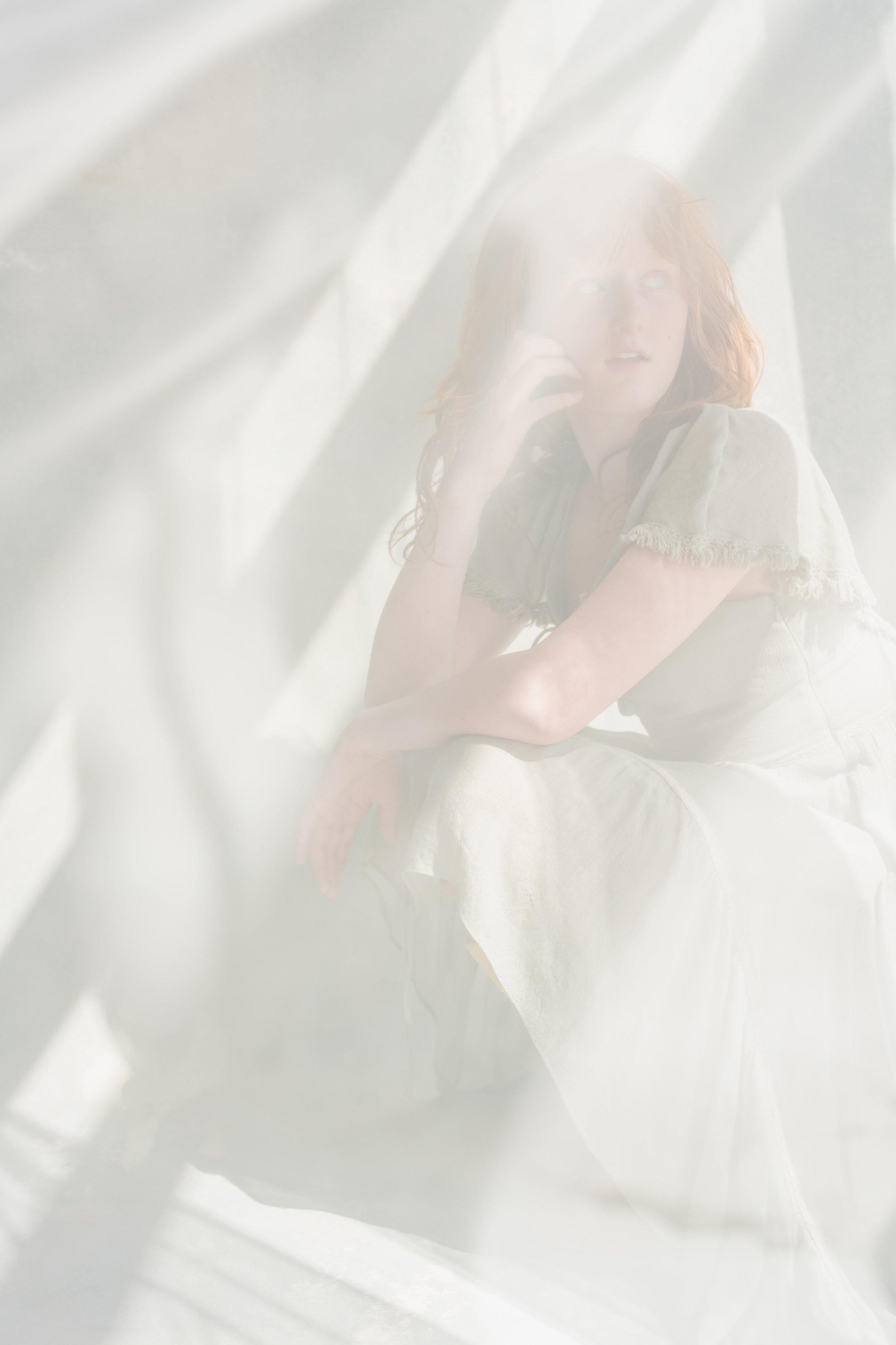 A young woman with red hair sitting on the ground, wearing a light-colored, frayed-sleeve dress, looking up with a surprised or curious expression, surrounded by soft, diffused light and flowing white fabric.