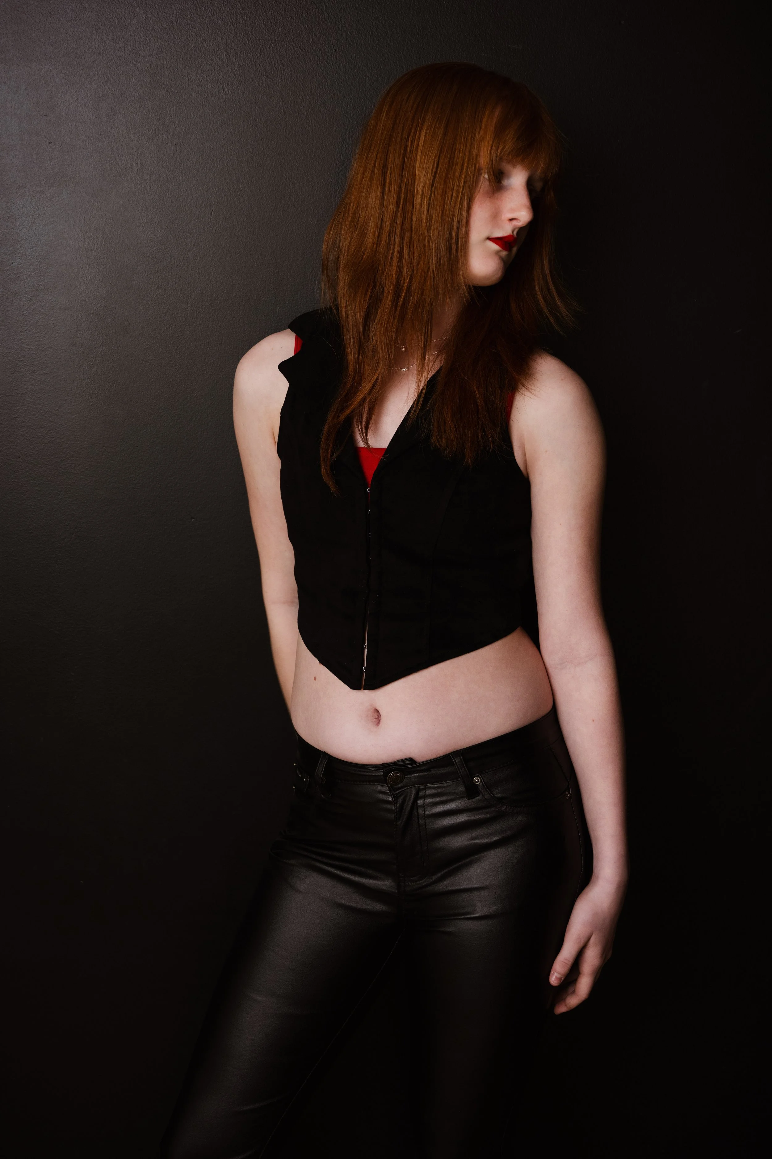 A woman with long red hair, wearing a black sleeveless top and black leather pants, standing against a black background with her head slightly turned to the right.