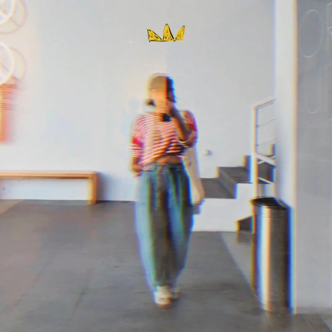 A woman in a striped shirt and loose pants takes a mirror selfie in a room with minimal decor, a small yellow crown drawing on the wall above her head.