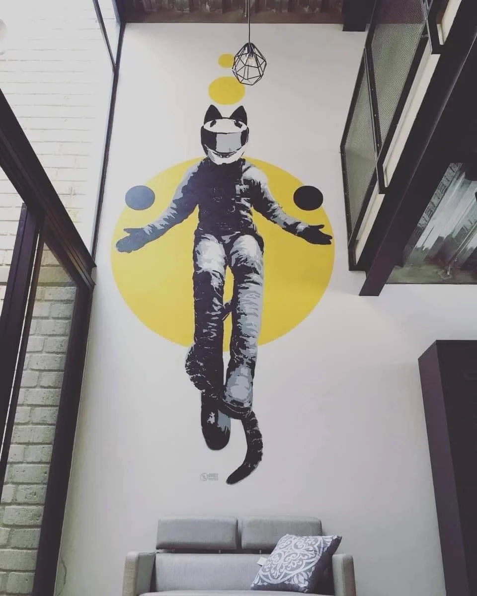 Wall art depicts a cat-human hybrid figure with a smiling cat mask, floating in front of a yellow circle and smaller colorful circles, with a black geometric ceiling light hanging above.